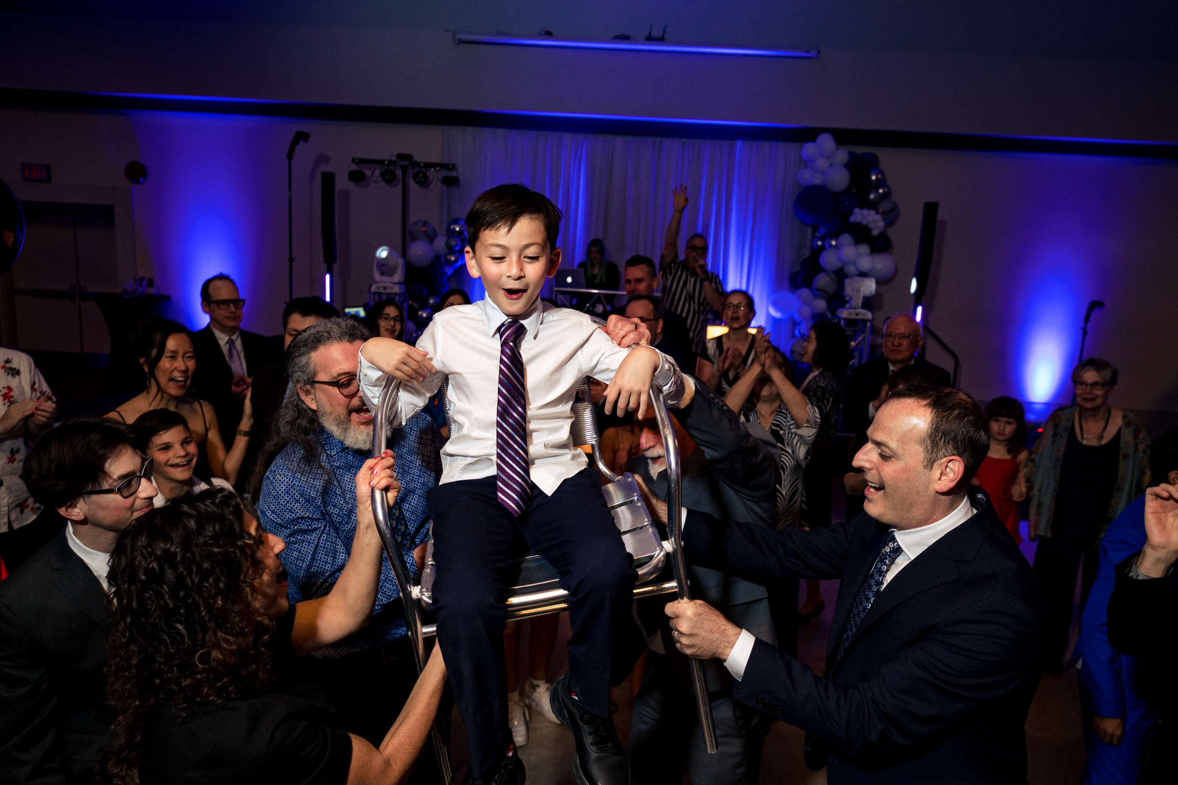 A young boy dressed in a white shirt and tie is being lifted on a chair by four adults during a celebration. The room is filled with people cheering and clapping, and is lit with blue lighting. In the background, there are balloons and party decorations.