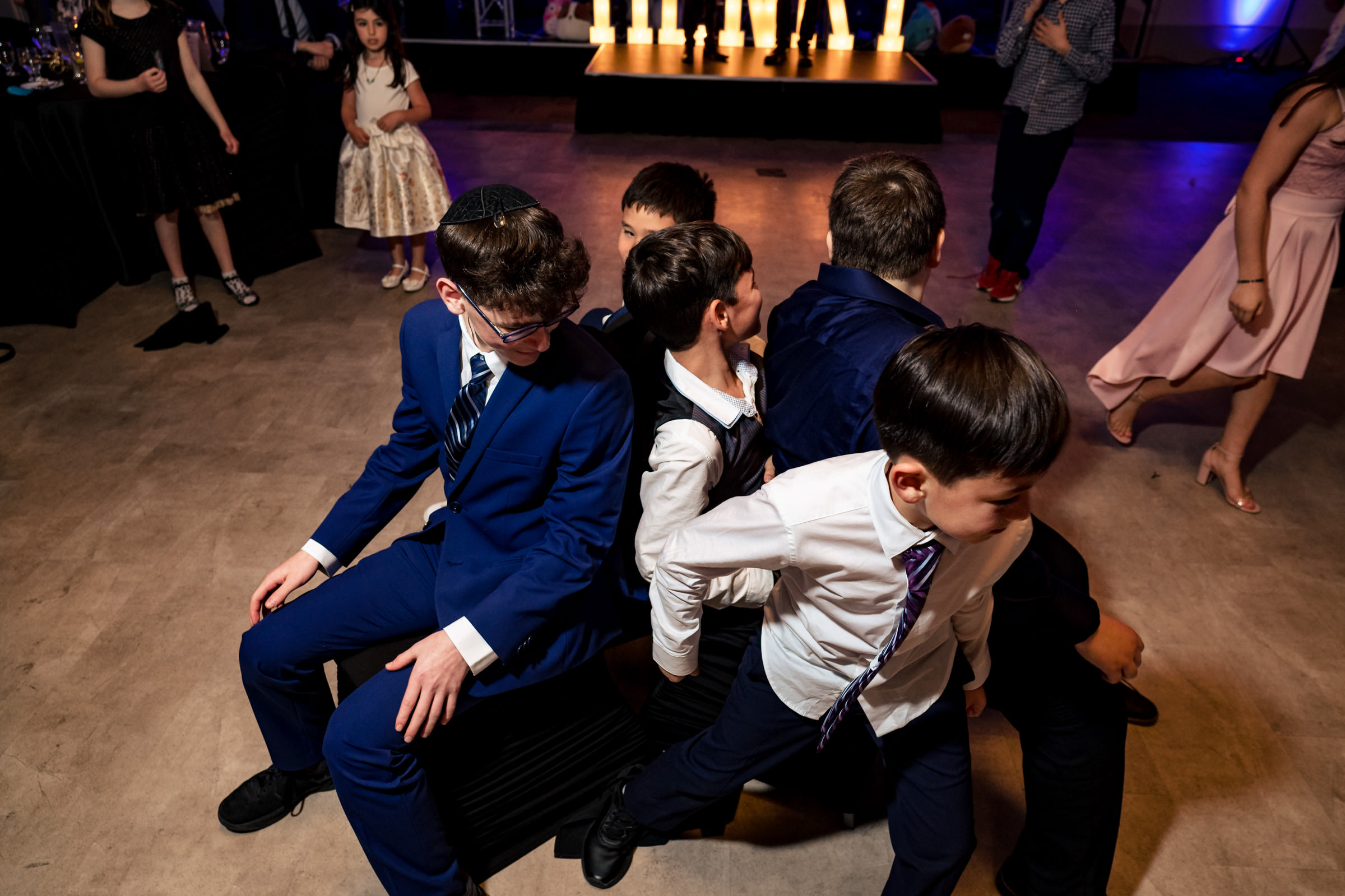 A group of boys dressed in formal attire play a game of musical chairs at a celebration. They are sitting back-to-back on the chairs, and one boy in a blue suit is looking over his shoulder. Some people can be seen standing and watching in the background.