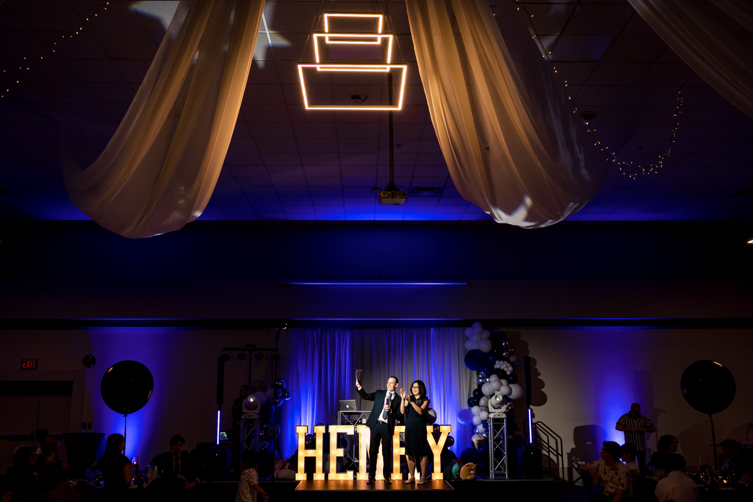 A stage is lit up with the name "HENLEY" in large illuminated letters. Two people are standing on the stage, with one speaking into a microphone. The ceiling is adorned with draped white fabric and hanging lights, adding a decorative touch to the banquet hall ambiance.