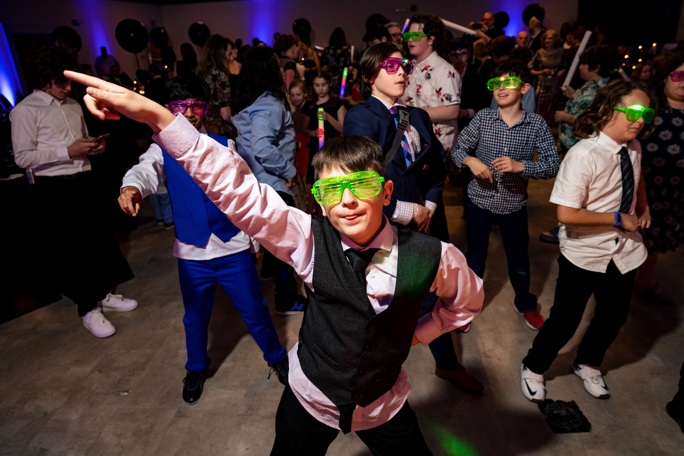 A group of children are dancing enthusiastically at a party. The boy in the foreground is striking a pose with his arm pointed upwards, wearing a vest, tie, and green light-up glasses. Other children in the background also wear light-up glasses and colorful outfits.
