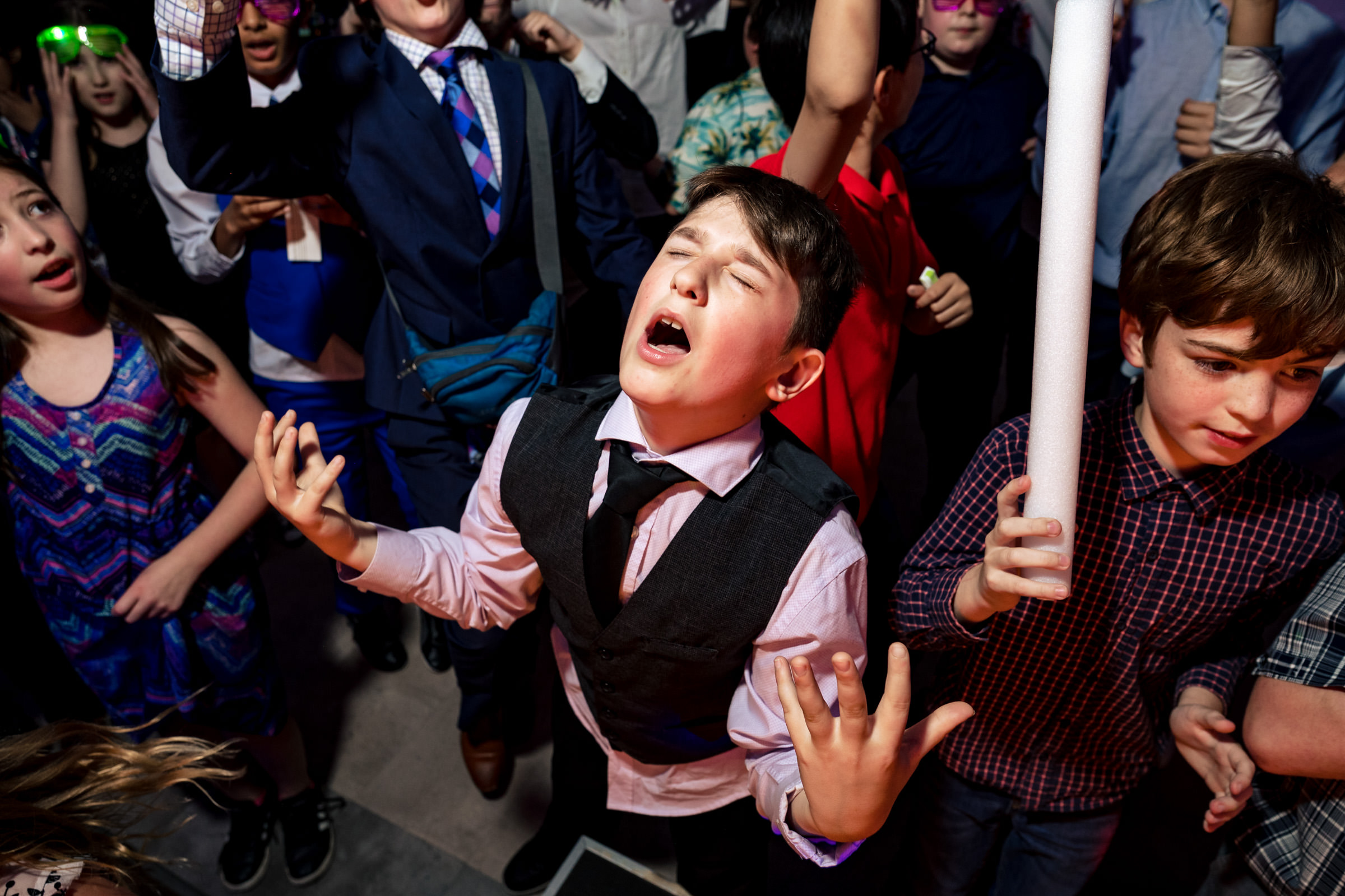 A young boy dressed in a pink shirt, black vest, and tie stands amidst a group of people, his eyes closed and his hands raised in an expressive gesture, possibly singing or passionately speaking. People around him are engaged, with some cheering and holding props.