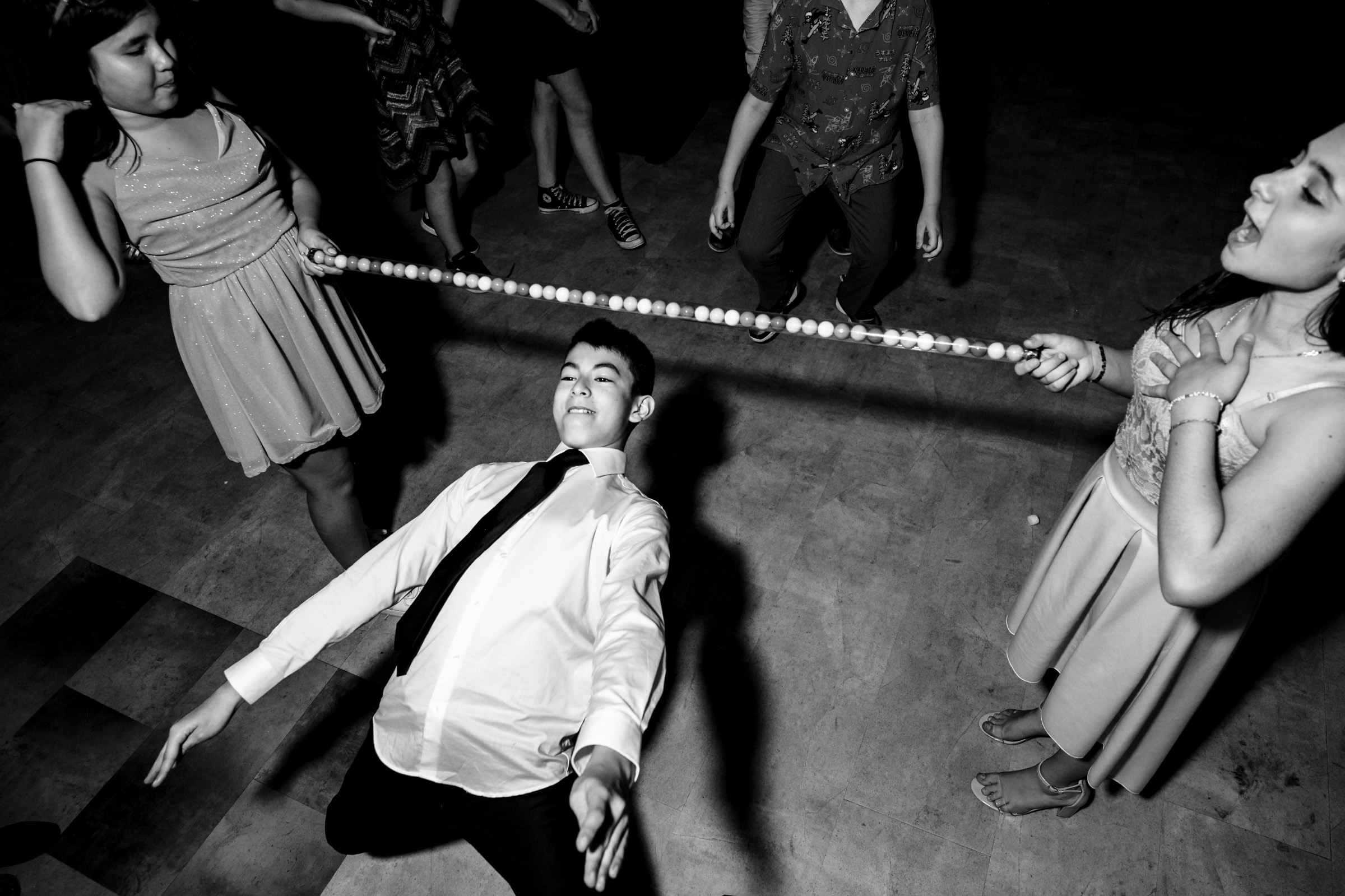 A black and white photo of a young man in a white shirt and tie leaning back to pass under a limbo stick held by two people. Several others stand nearby, watching and smiling. The scene appears to be festive and joyful.