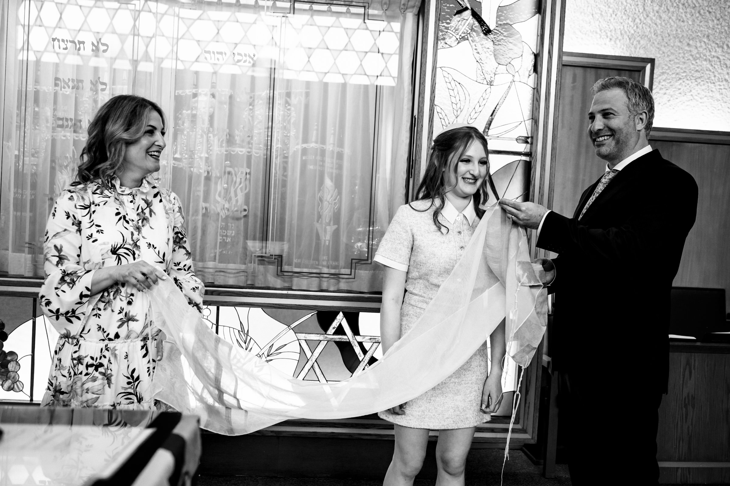 A black and white photo shows three people standing indoors: a smiling woman on the left, a happy girl in the center, and a smiling man on the right holding a sheer fabric over the girl's shoulders. They appear to be participating in a celebratory or religious event.
