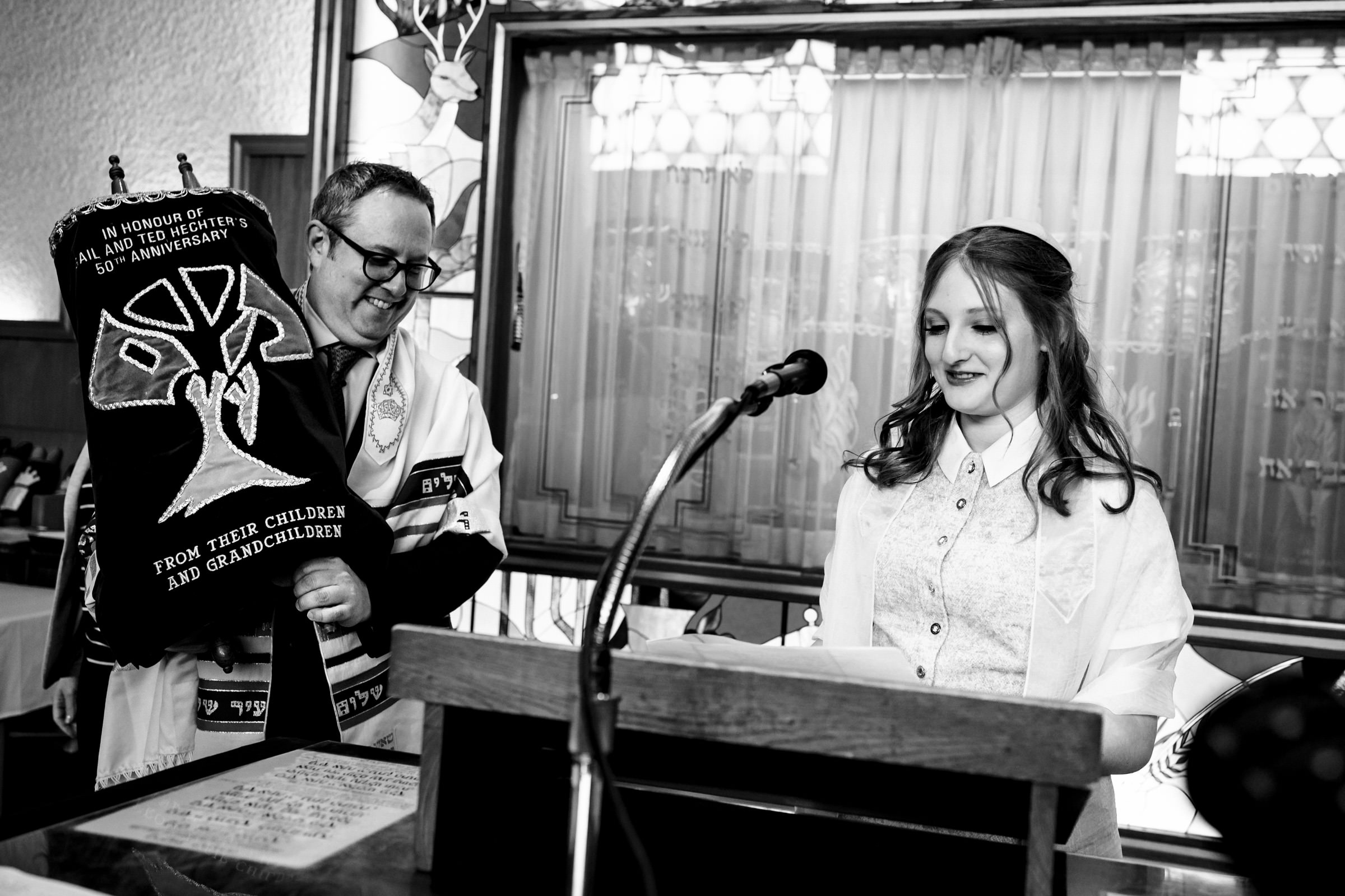 A black-and-white photo shows a woman speaking at a podium with a microphone, smiling. Beside her, a man wearing glasses and a prayer shawl holds a Torah scroll adorned with an embroidered design and text celebrating an anniversary from children and grandchildren.