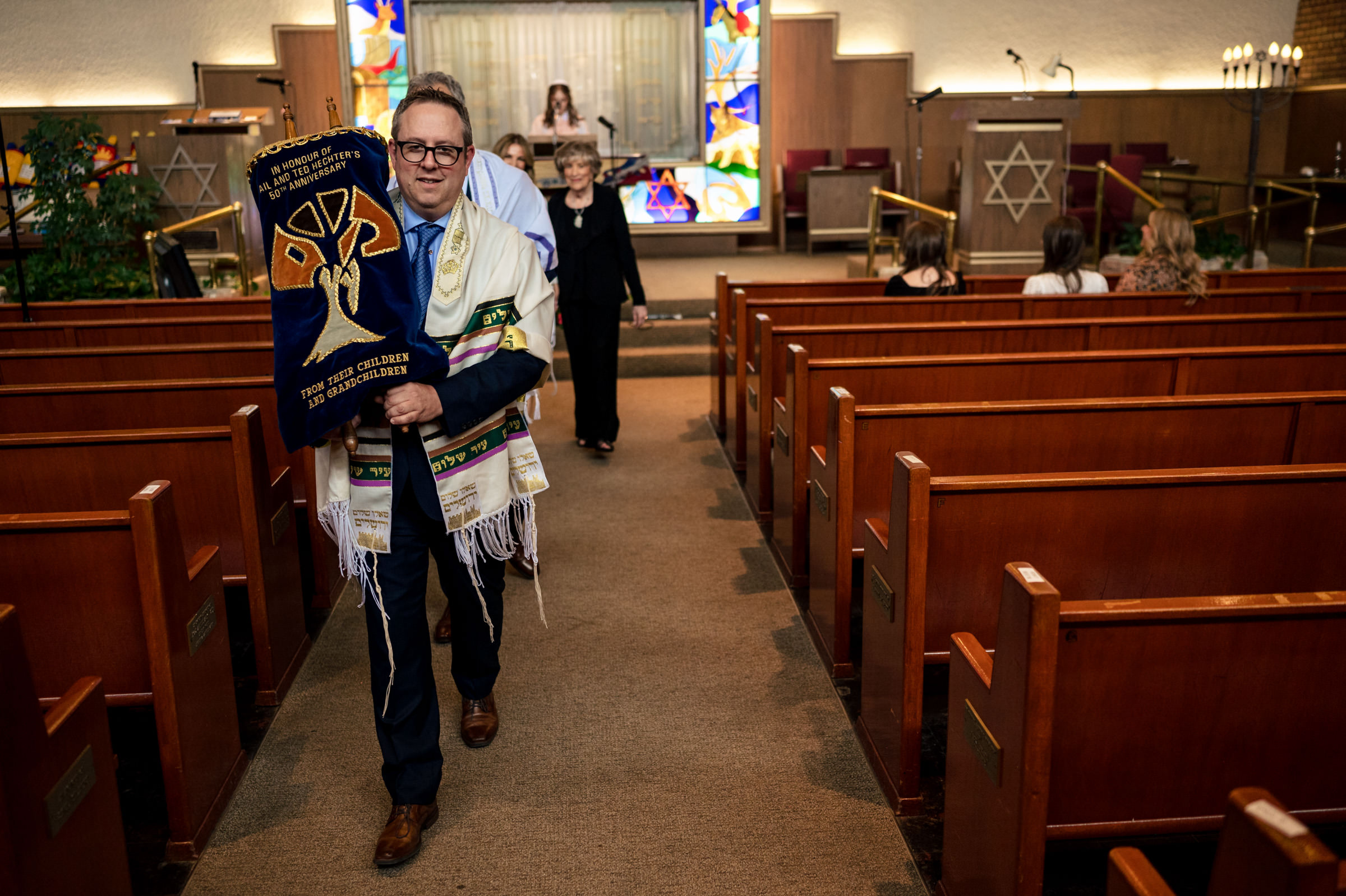A person walks down the aisle of a synagogue carrying a Torah scroll wrapped in blue fabric, followed by others. The synagogue is adorned with wooden pews and colorful stained glass windows. An individual stands at the bimah in the background.