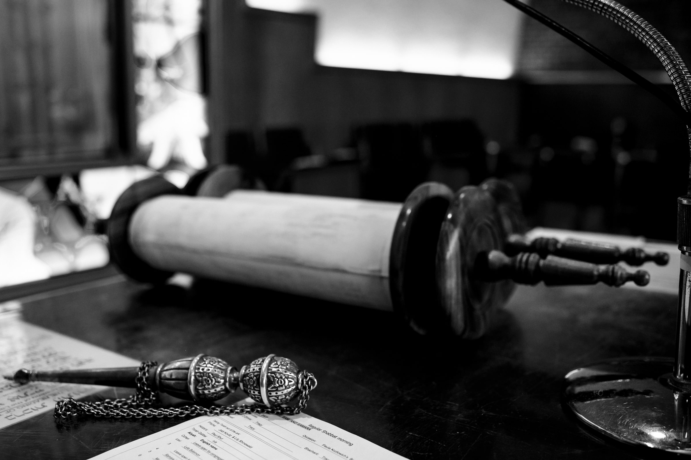 A black-and-white image of a Torah scroll placed on a table. The scroll is partially unrolled, revealing some text. In the foreground, there is a yad (Torah pointer) with intricate designs, and some papers. Blurred background shows chairs and a window.