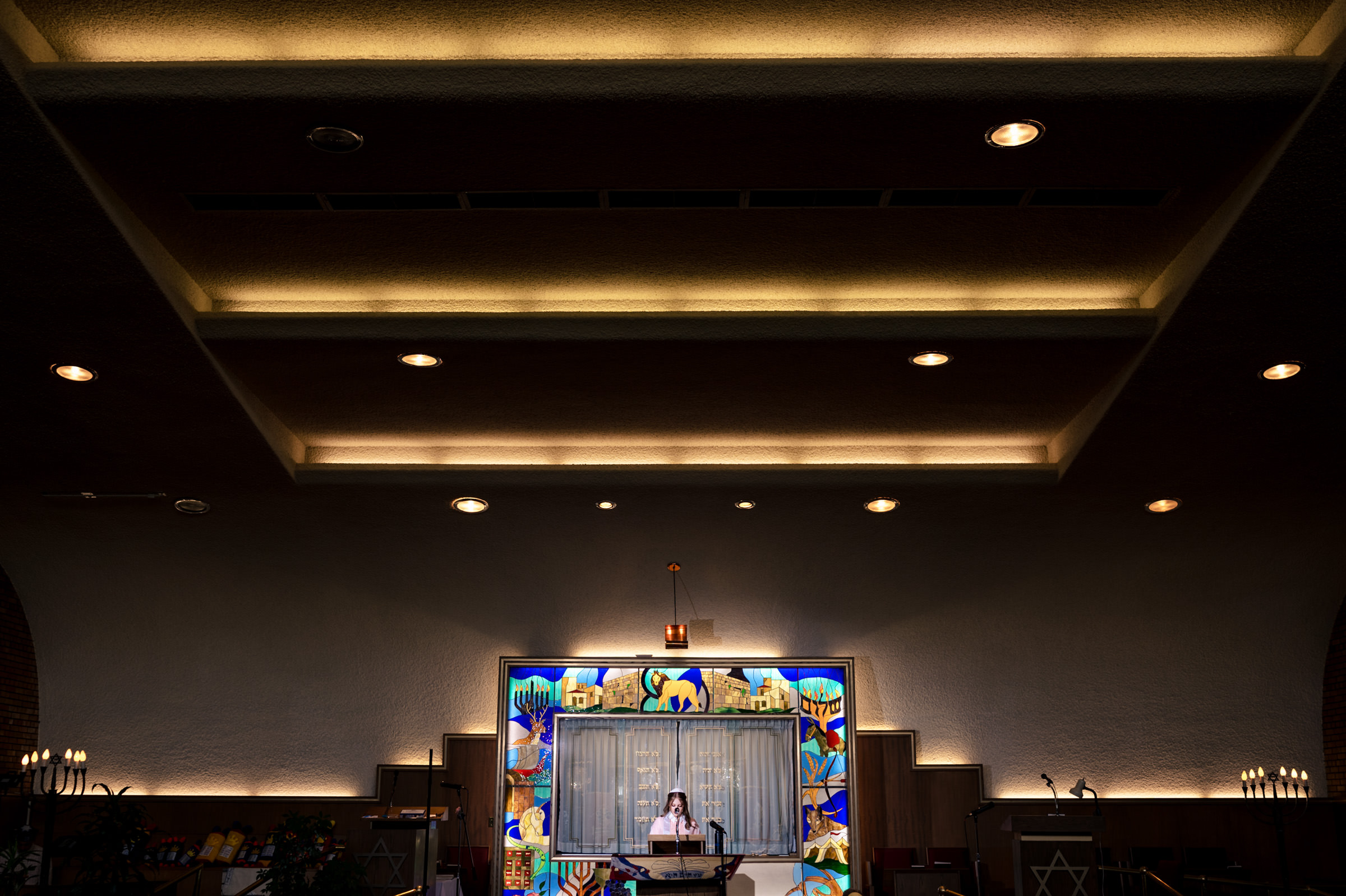 A person stands at the pulpit in a large, ornate synagogue. The ceiling has recessed lighting, and a colorful stained glass decorates the wall behind the pulpit.