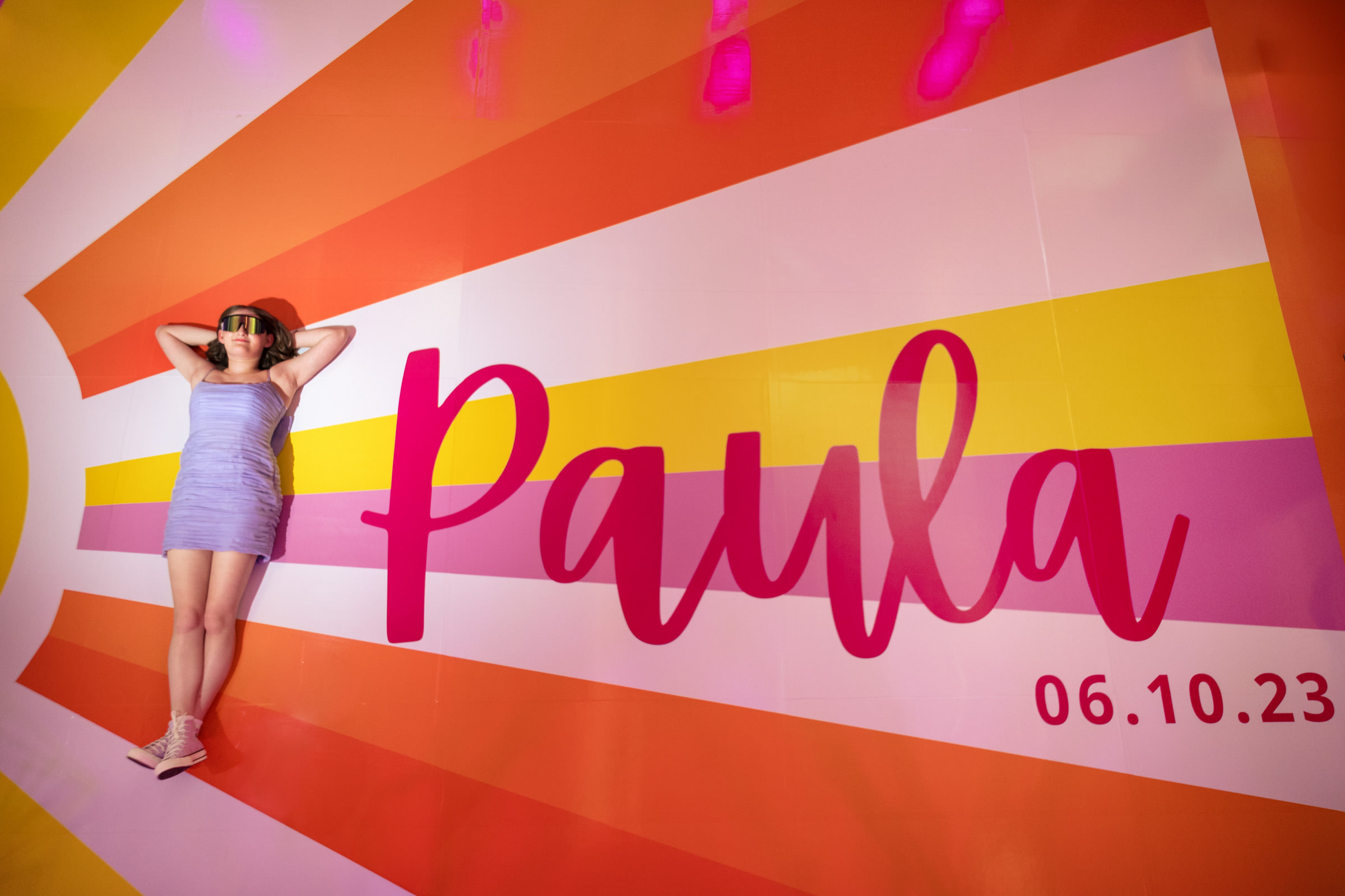A woman in a blue dress and sunglasses lounges against a brightly colored mural with rays of orange, pink, yellow, and white. The mural features the name "Paula" in large pink letters and the date "06.10.23." The lighting casts a vibrant glow.