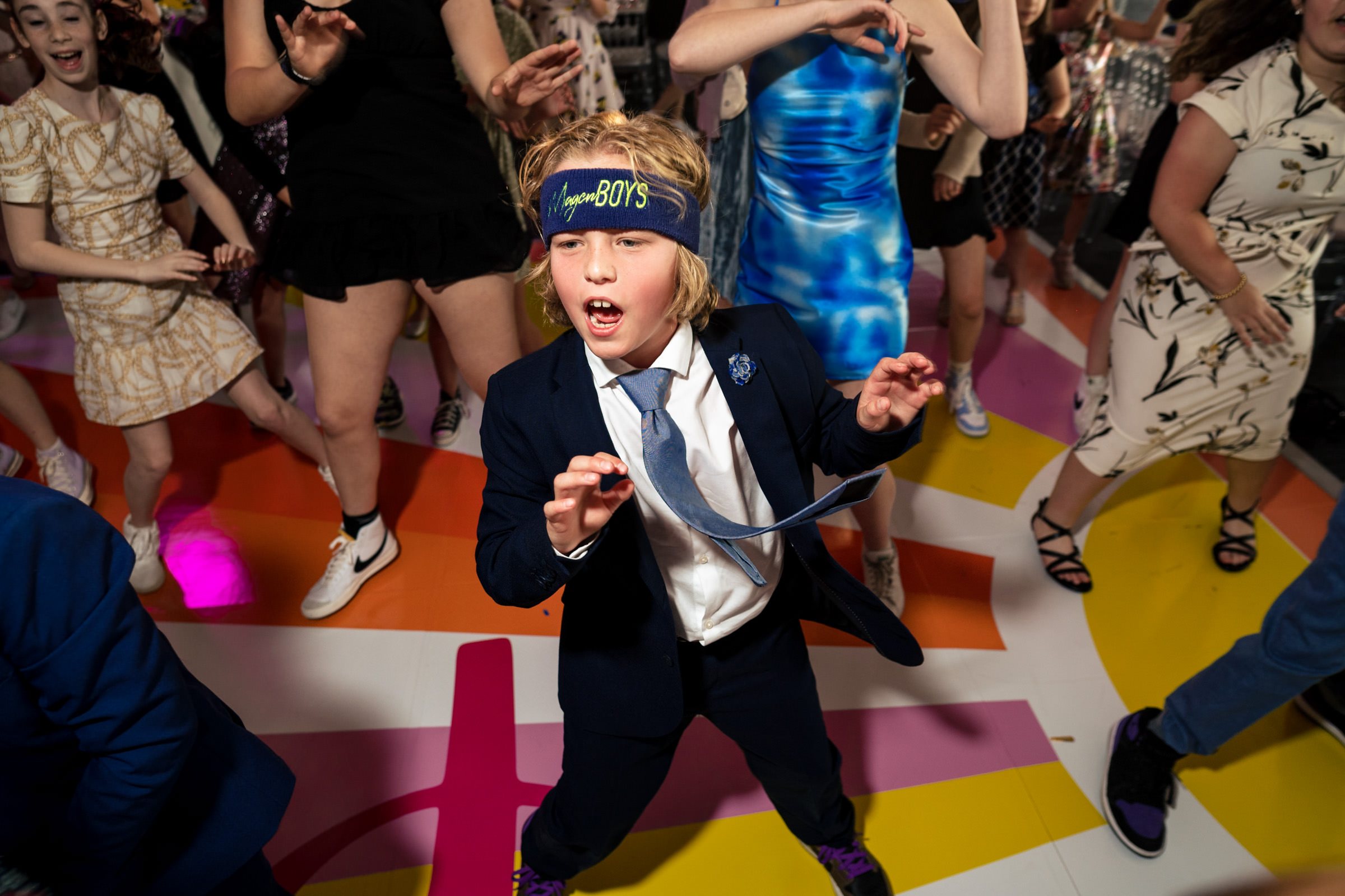 A young boy wearing a suit and tie, with a headband that reads "HappyBOYS," dances energetically among a group of people. The floor has colorful patterns, and others around him appear to be joining in the dance, creating a lively atmosphere.