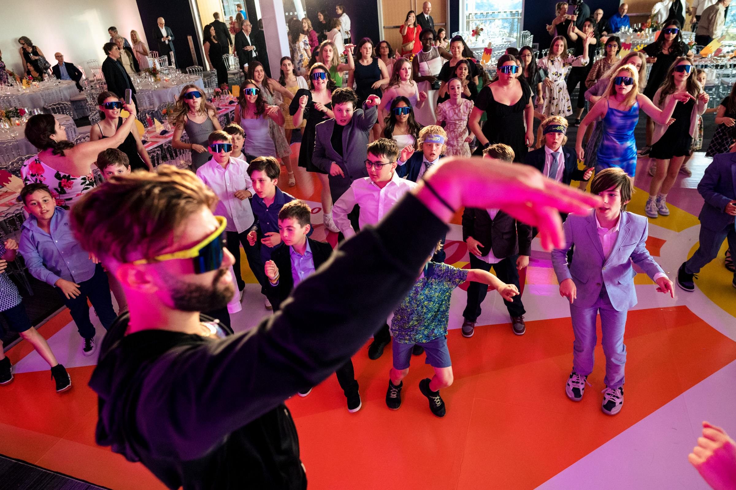 A lively dance party with people wearing colorful sunglasses under vibrant lighting. A man in the foreground gestures energetically while the crowd, including children and adults in various outfits, dances enthusiastically behind him. Tables with decorations are visible in the background.