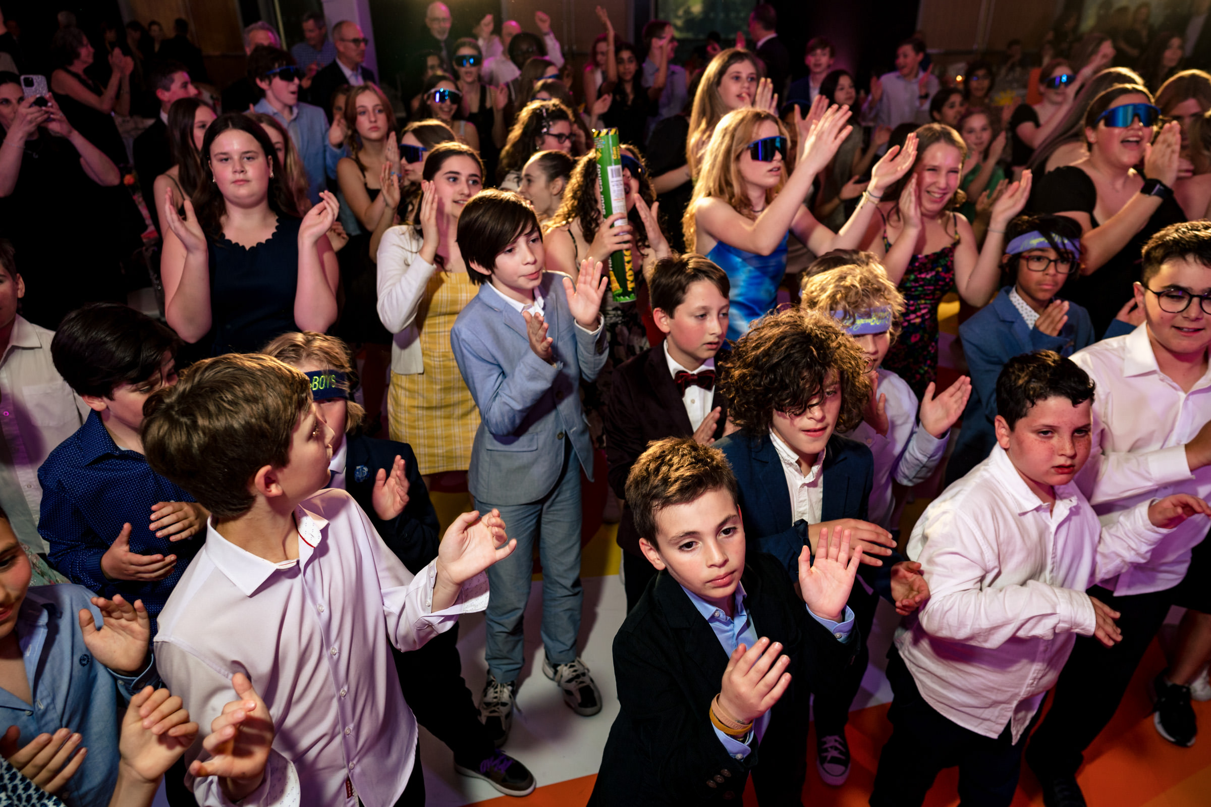 A lively crowd of young people, dressed in various formal and casual outfits, clap and dance together at an indoor event. Many are smiling and enjoying the music with some wearing vibrant accessories like sunglasses and headbands. The background shows more participants.