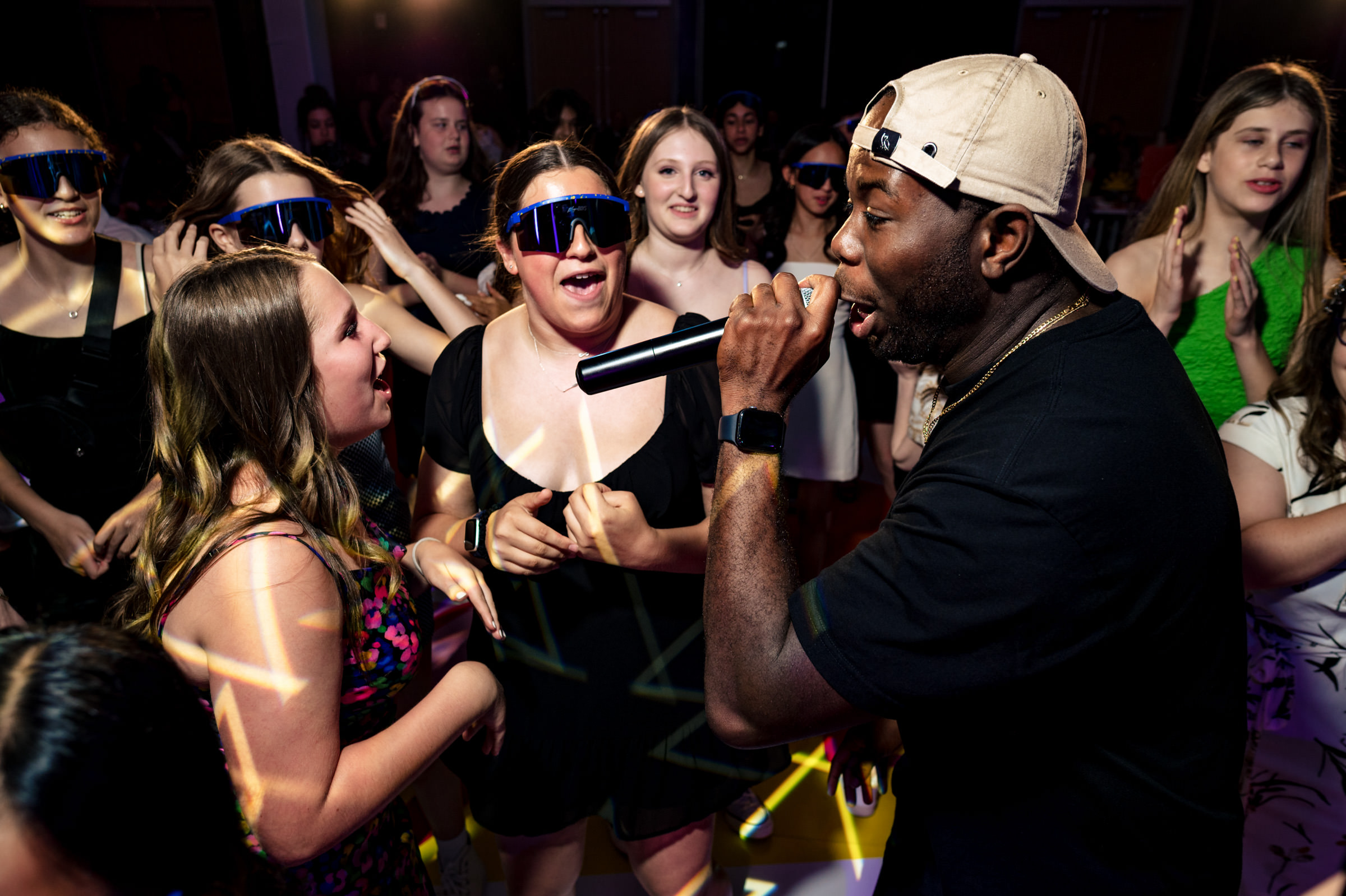 A performer wearing a cap and black shirt sings into a microphone while a group of enthusiastic women in colorful outfits dance and cheer around him. One woman near the front is holding her sunglasses up and smiling. The background is lively with multicolored lights.