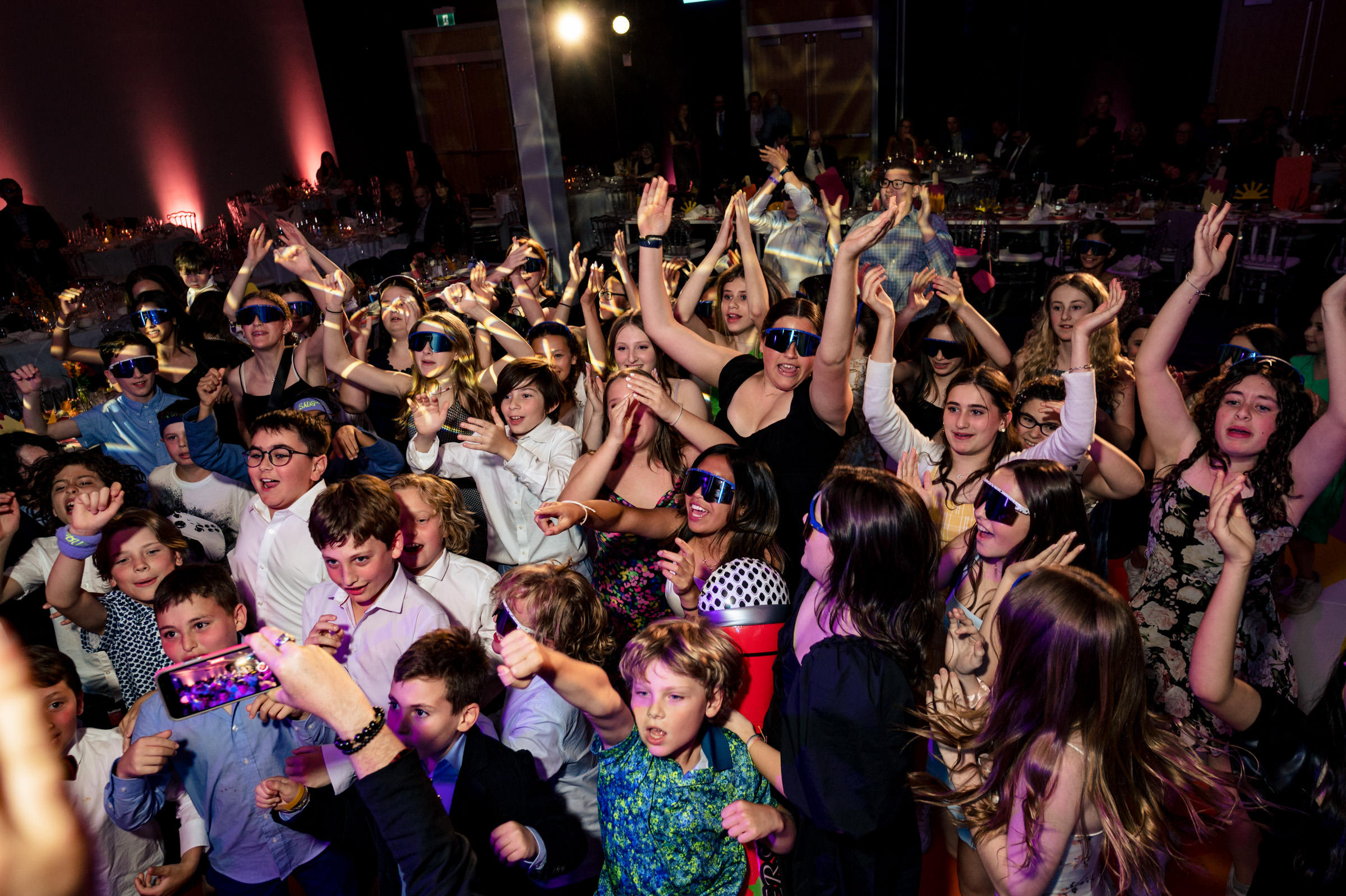 A lively party scene with a large group of people dancing and cheering under colorful lighting. Many are wearing sunglasses and festive outfits, and some are raising their hands in excitement. The atmosphere is joyous and energetic.