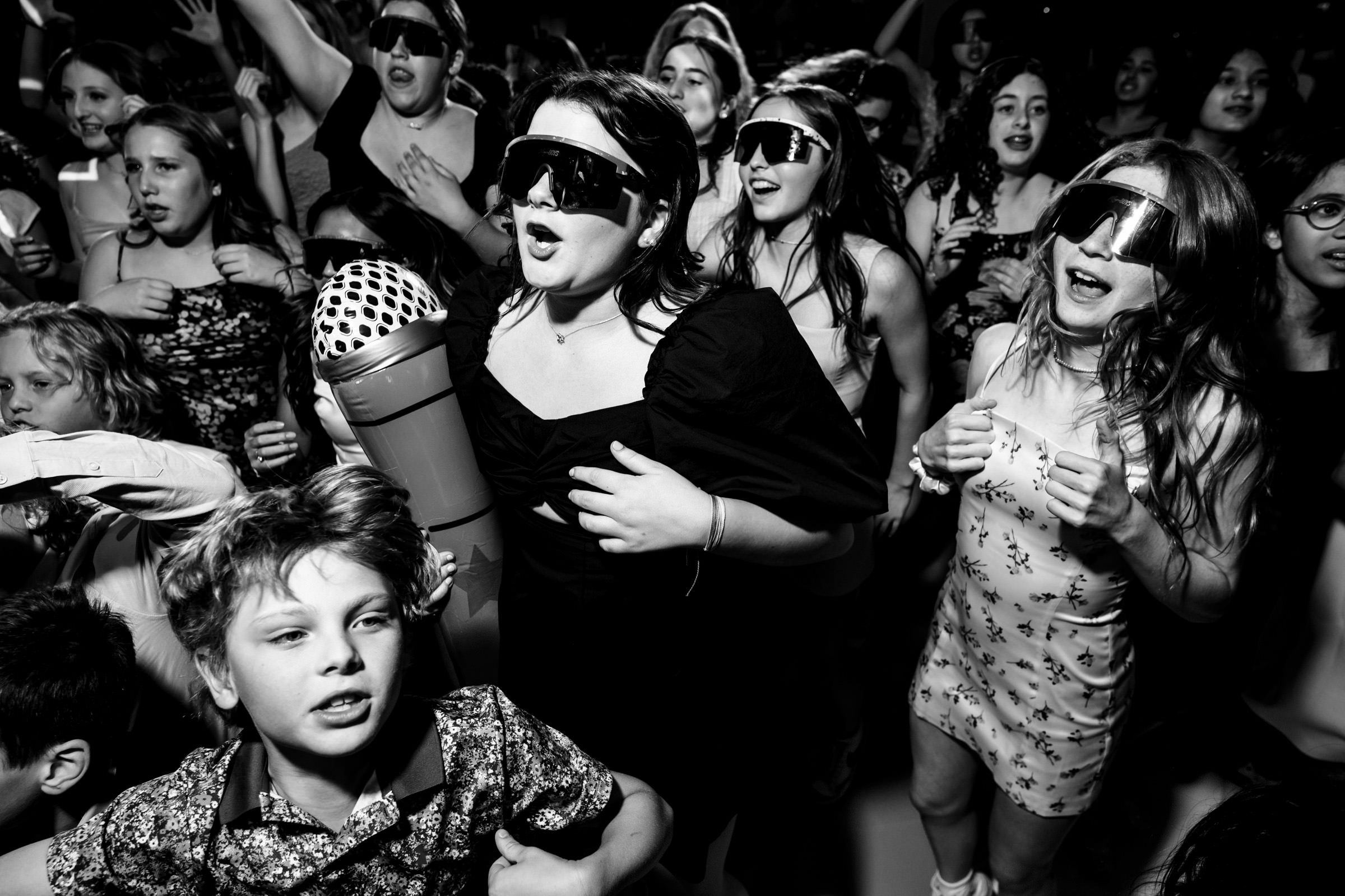 A group of children are energetically dancing and singing while wearing large, futuristic sunglasses indoors. One child in the front holds a large inflatable microphone. The scene is lively and filled with joyful expressions. The image is in black and white.