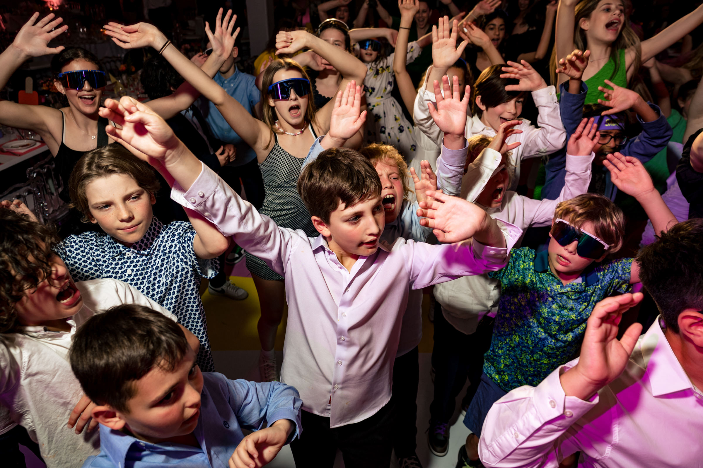 A lively group of children and teenagers dance energetically with their hands in the air. Some wear sunglasses and colorful attire, while others are in white shirts. The scene is vibrant, with everyone enjoying the music and the festive atmosphere.
