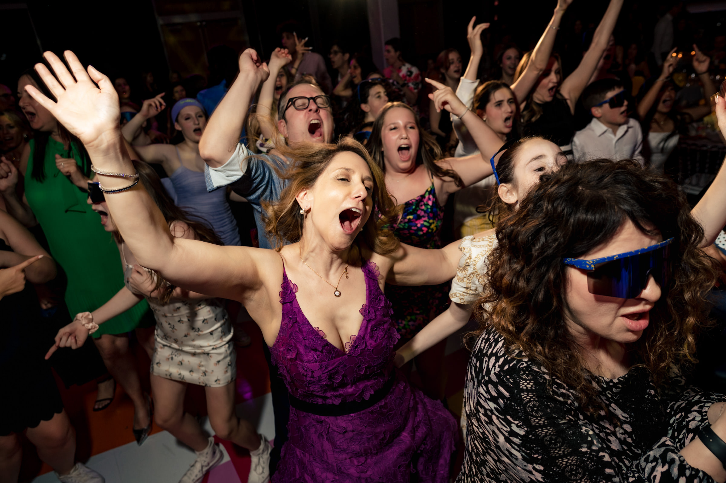 A crowd of people energetically dances and sings with their hands in the air at a lively party. The woman in the foreground wears a purple dress, and another woman beside her sports large, blue sunglasses. The atmosphere is jubilant and enthusiastic.