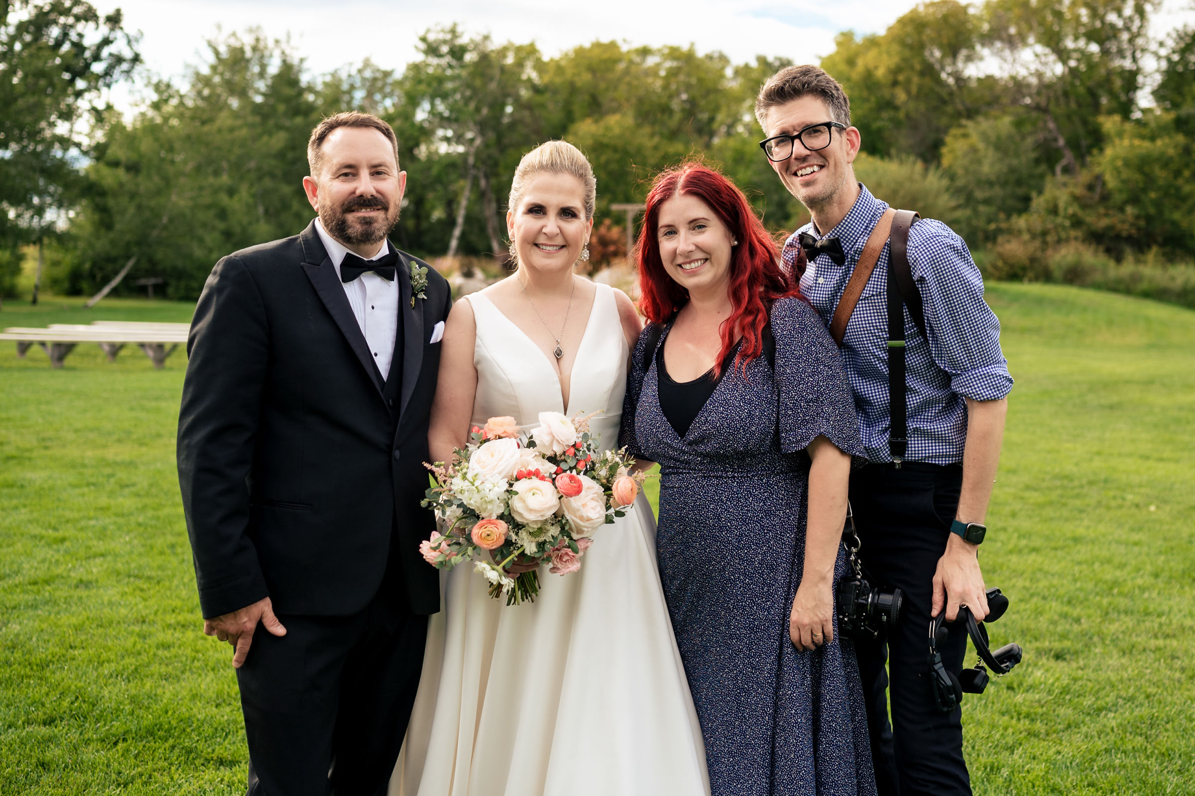 A bride and groom pose outdoors with two other people. The bride wears a white dress and holds a bouquet, while the groom dons a black tuxedo. The two others stand beside them; the woman wears a blue dress and the man has a camera slung over his shoulder.