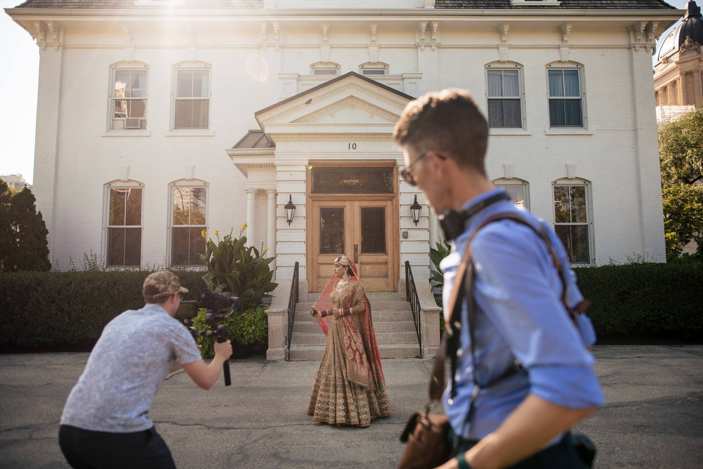 A bride in a traditional wedding dress and veil stands in front of a large white building, posing for a videographer. Another person with camera equipment is in the foreground, partly facing away from the camera. The sun shines brightly, casting soft light.