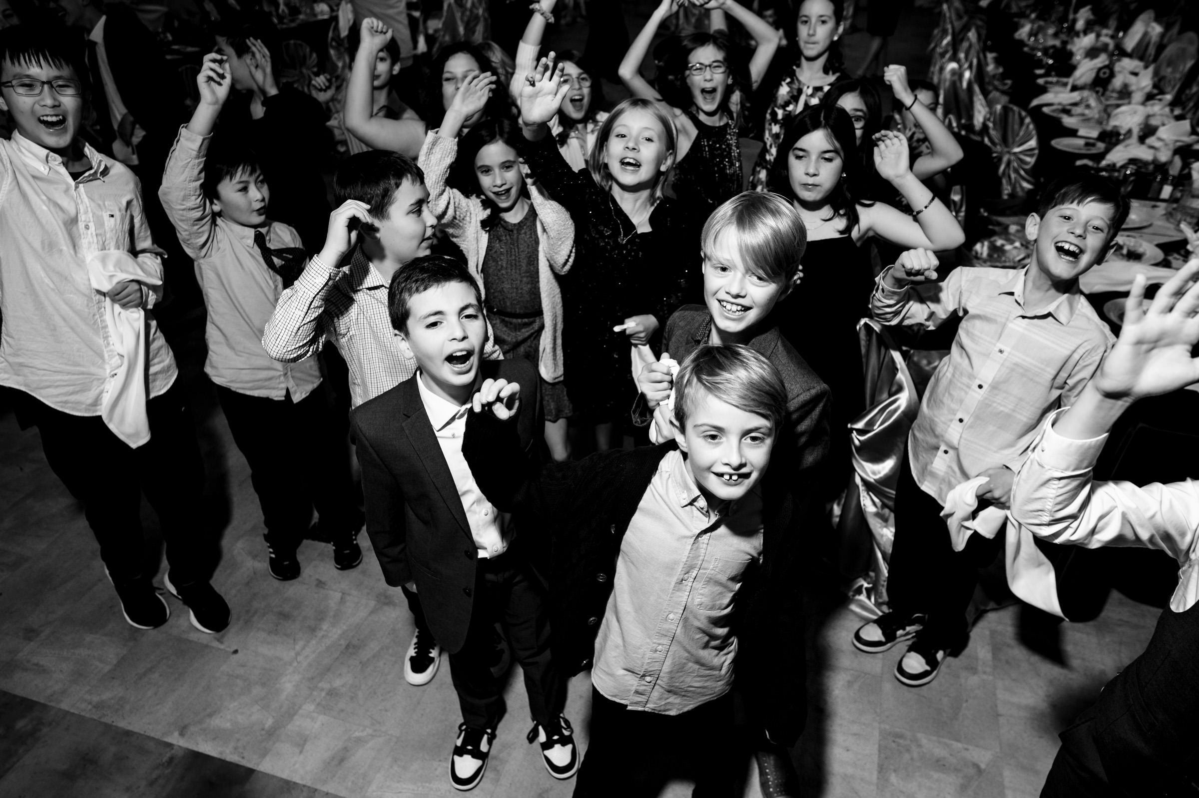 A group of children cheerfully pose with their hands up at what appears to be a party or celebration. They are dressed in semi-formal clothing and are closely gathered together, smiling and looking at the camera. The background shows other guests and tables.