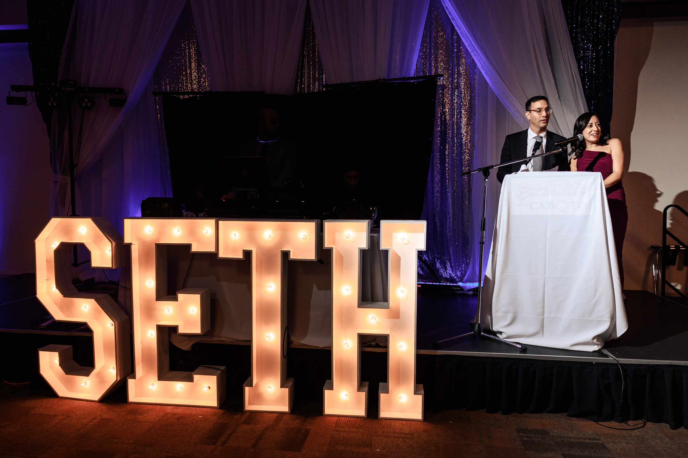 A man and a woman stand at a podium on a stage, speaking into microphones. Behind them, large illuminated letters spell out "SETH" against a backdrop of draped fabric and sparkly decorations. The scene appears to be at an event or celebration.