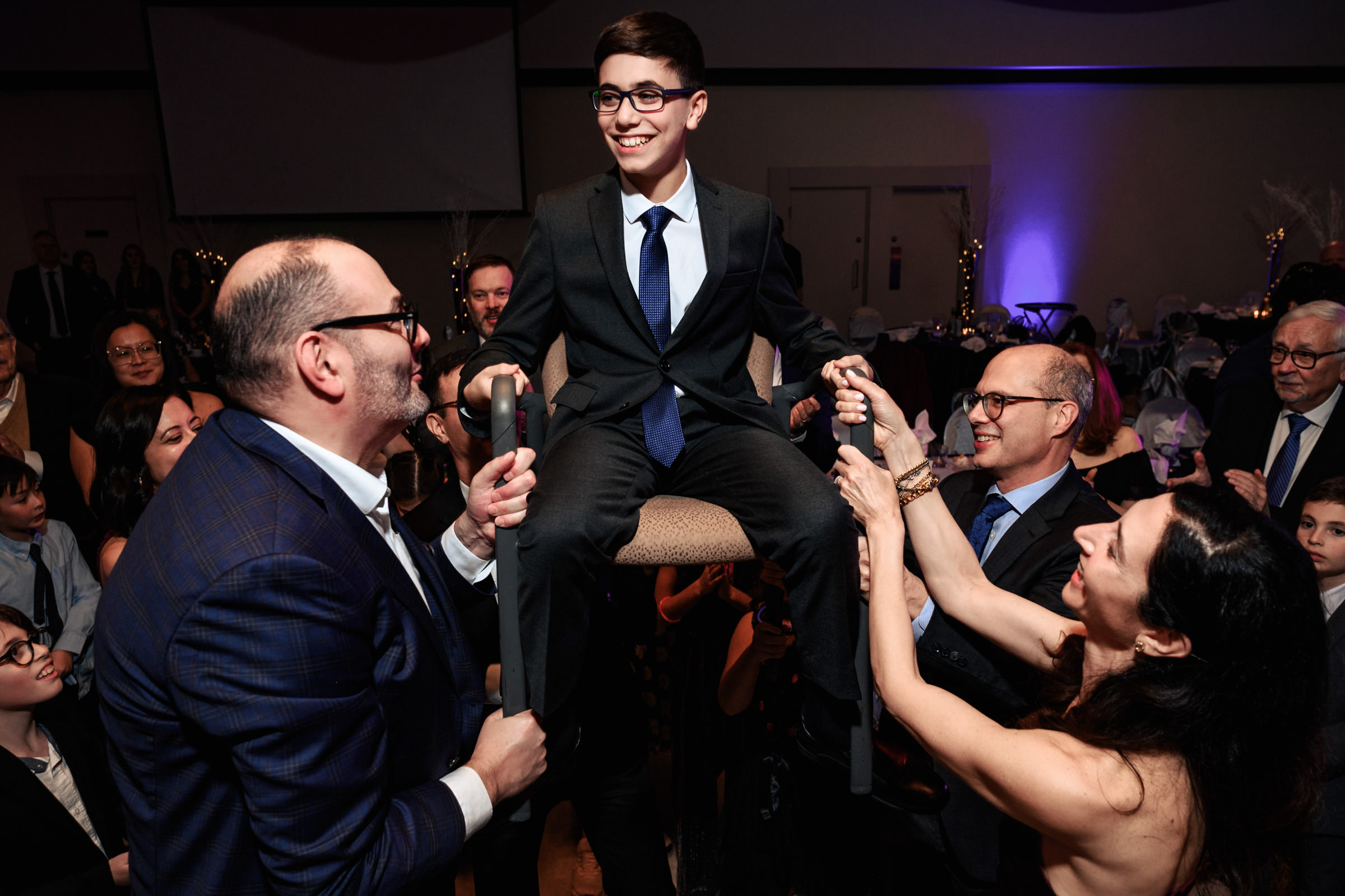 A young boy in a suit and glasses is lifted on a chair by three adults during a celebration, smiling and holding onto the chair arms. The background shows dim lighting with guests and tables, suggesting a joyous event or party.