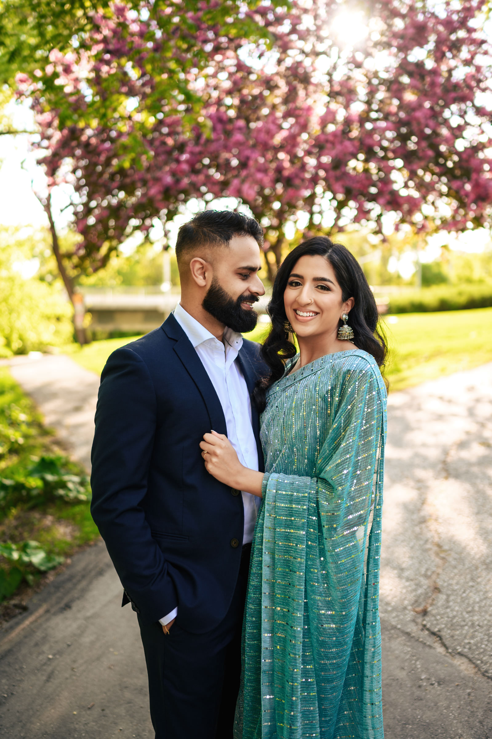 A couple smiling and standing close to each other on a sunny day during their Indian engagement shoot. The man is wearing a dark suit, and the woman is dressed in a sparkling green traditional outfit. They are outdoors with a vibrant, blossoming tree in the background.