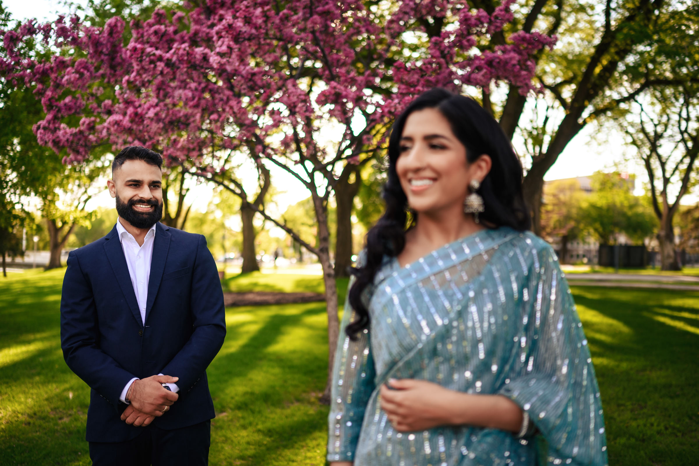 A man and a woman are outdoors in a park with blooming trees and green grass. The man, wearing a dark suit, stands in the background smiling. The woman in the foreground is dressed in a sparkly teal saree, her face slightly out of focus, capturing the essence of an Indian engagement shoot.