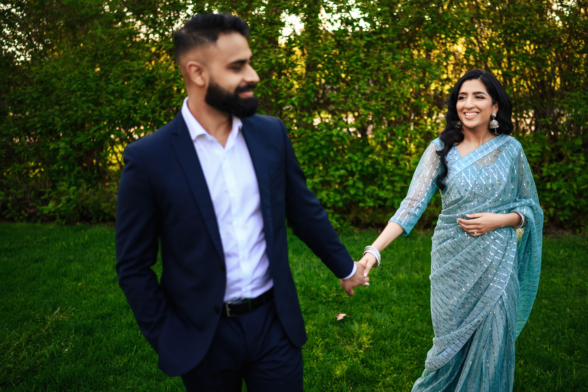 A man in a navy blue suit holds hands with a woman wearing a light blue sari with silver embellishments. They are walking on green grass with lush foliage in the background. The woman is smiling, and both appear to be in a cheerful mood, capturing the joy of their Indian engagement shoot.