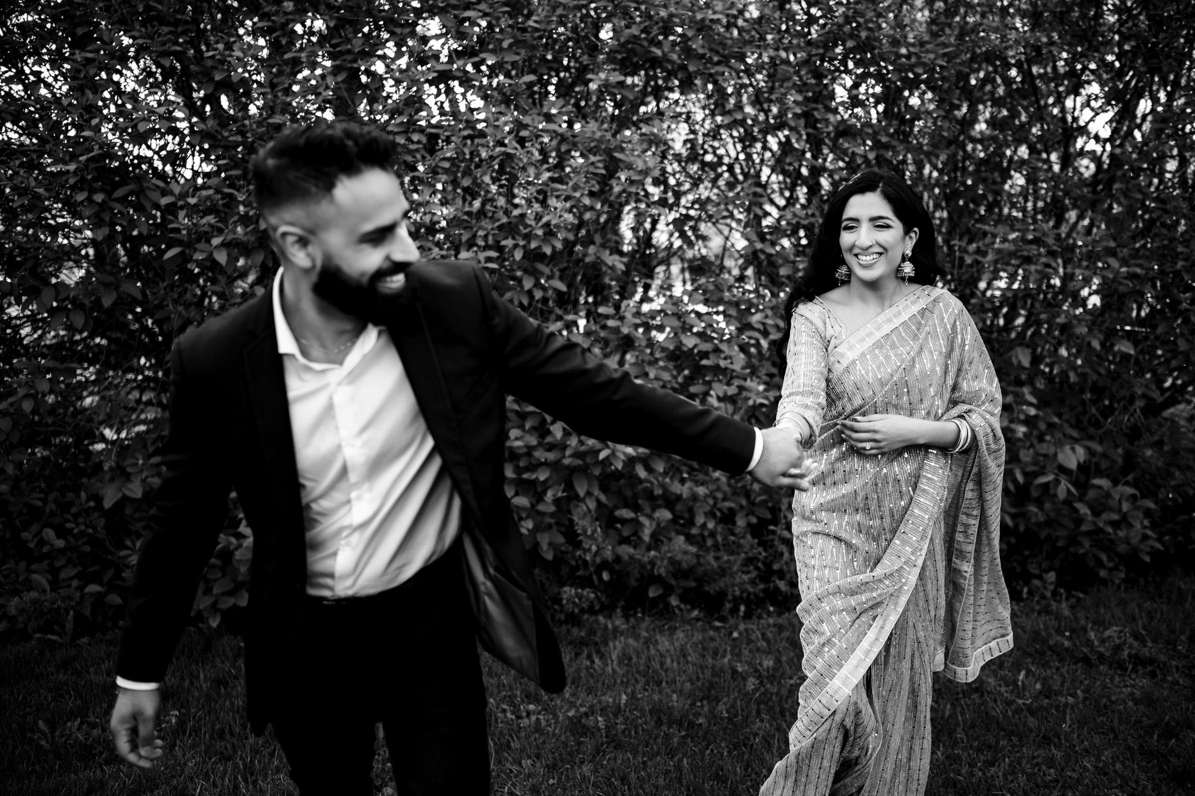 A black and white image of a couple holding hands outdoors during their Indian engagement shoot. The man, in a suit, is in front, looking back and smiling. The woman, in a traditional saree, is behind him, also smiling. They are surrounded by dense foliage.