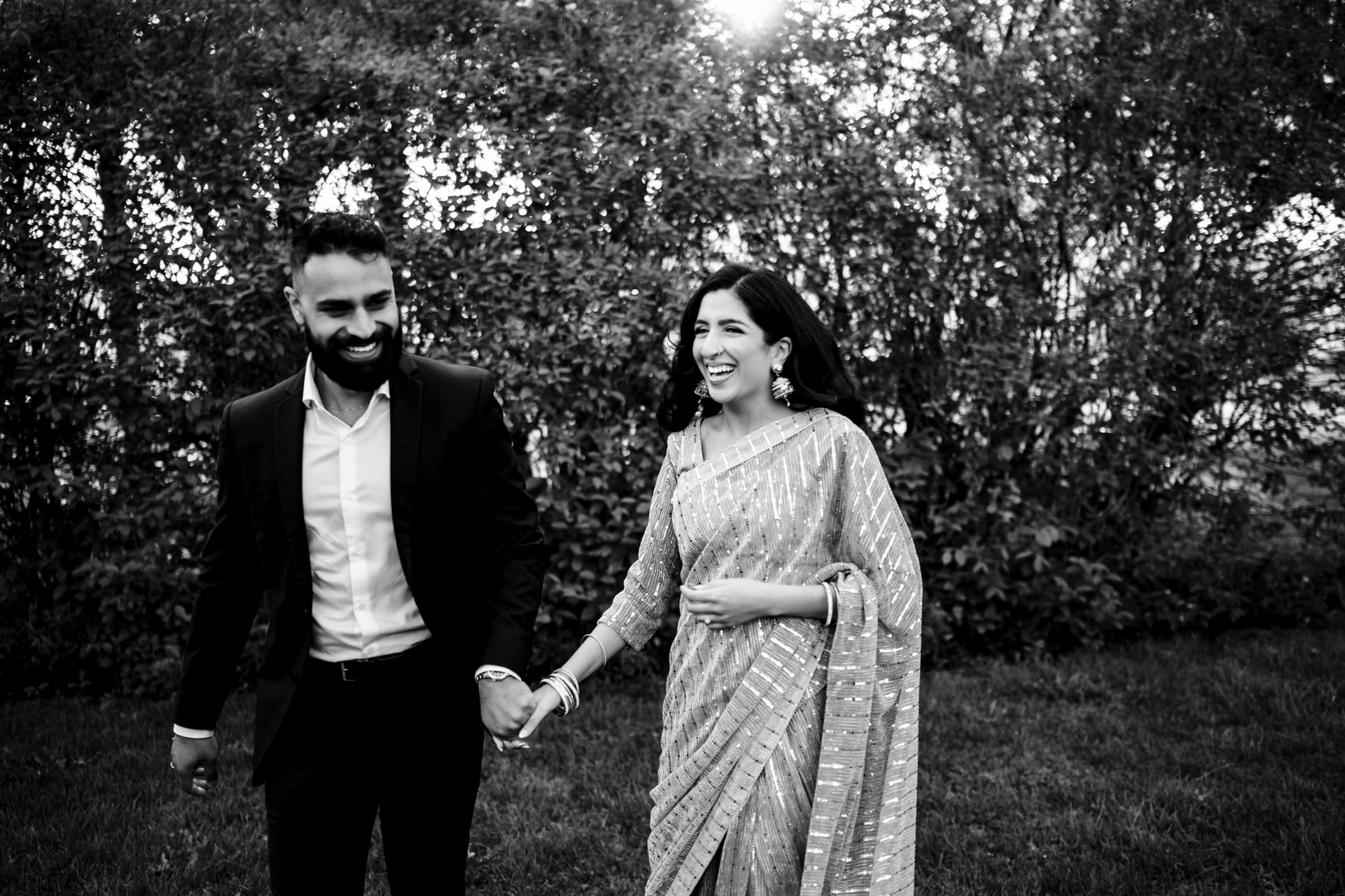 A black and white photo of a couple walking hand in hand outdoors during their Indian engagement shoot. The man, wearing a dark suit, smiles while looking at the woman, dressed in a patterned saree and smiling back at him. They are surrounded by lush greenery.