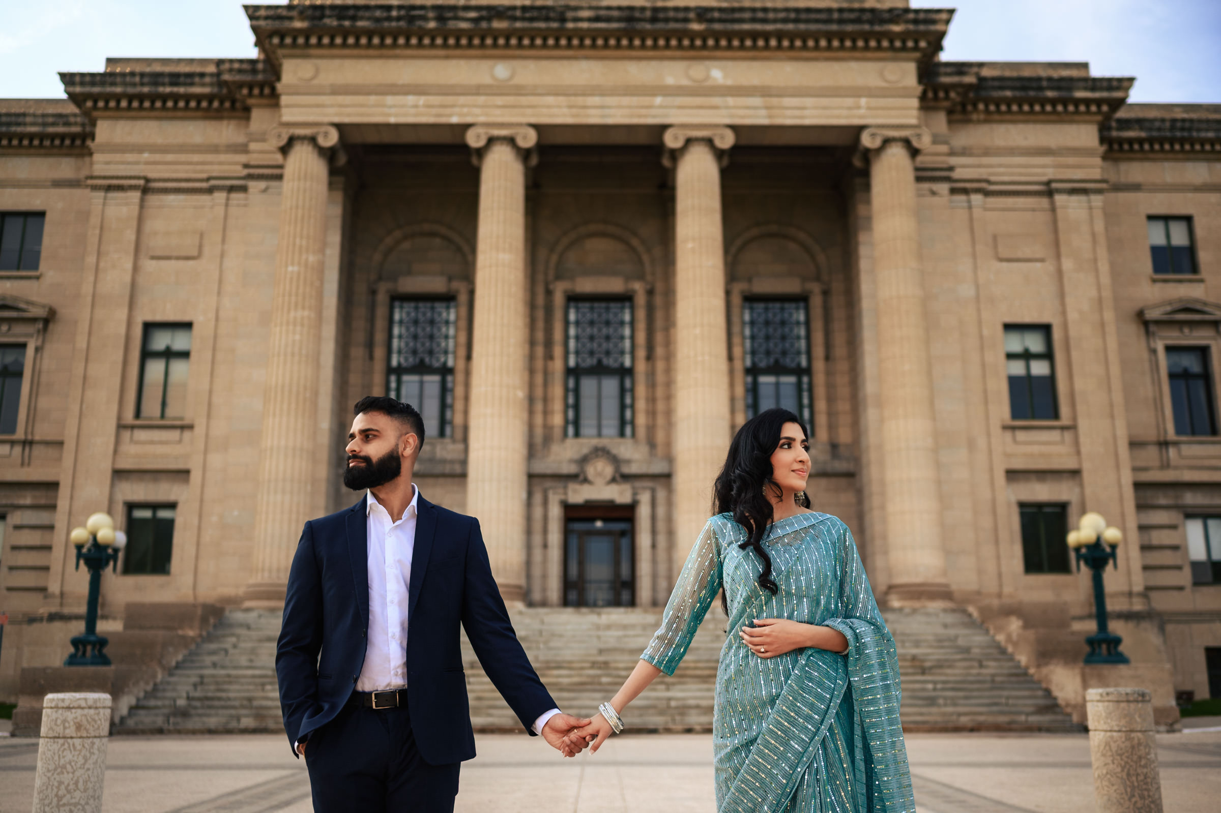 A man and woman holding hands stand in front of a grand building with large columns and steps, capturing their Indian engagement shoot. The man is wearing a dark suit, while the woman is dressed in a traditional, teal-colored outfit. Both are looking in opposite directions.