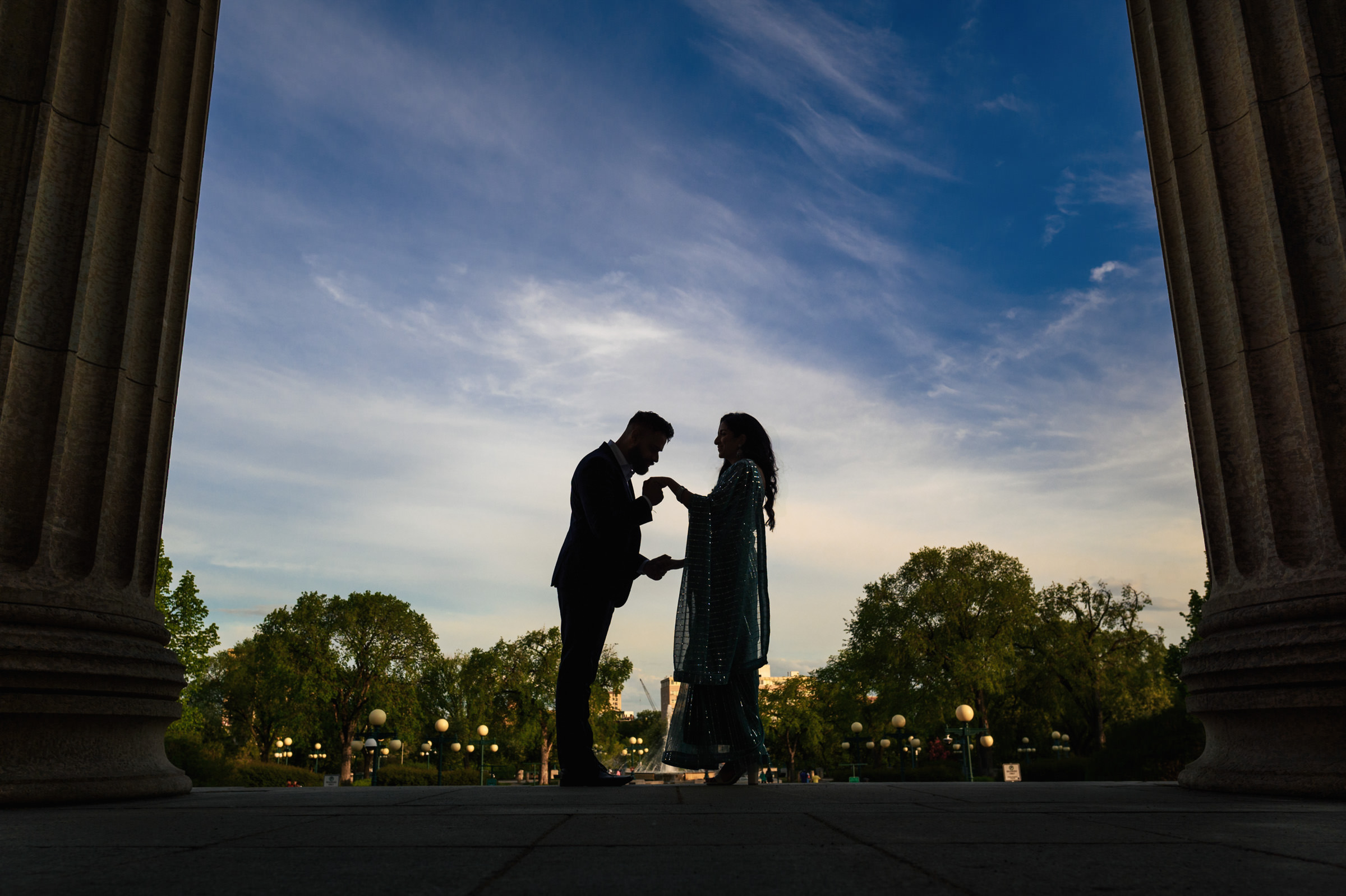 Silhouetted couple in formal attire stand between two large columns, holding hands and looking at each other. The background, evocative of an Indian Engagement Shoot, features a clear blue sky with some clouds, greenery, and distant street lamps, suggesting an outdoor setting at dusk.