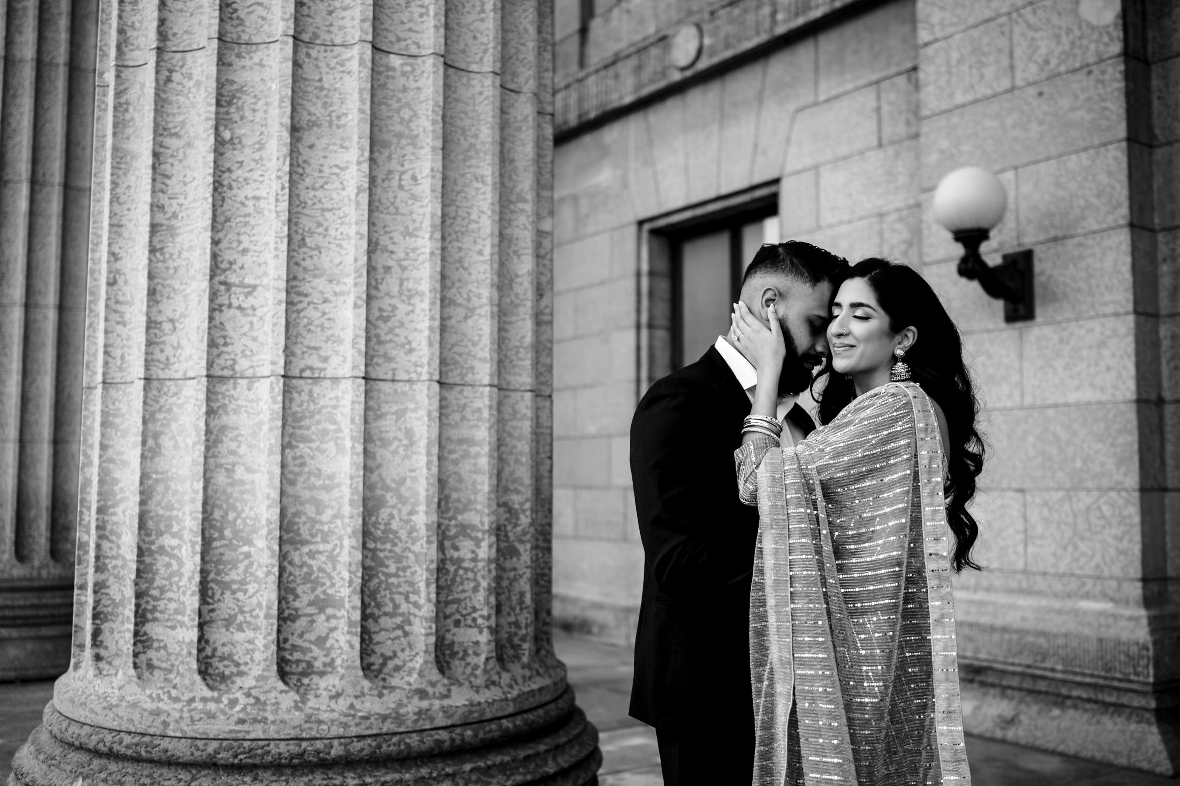 A couple stands close during their Indian engagement shoot, with the man gently cupping the woman's face. They are outside a stone building with large pillars. The woman wears a traditional, shimmering sari, and they share an intimate moment, both with serene expressions. The photo is in black and white.