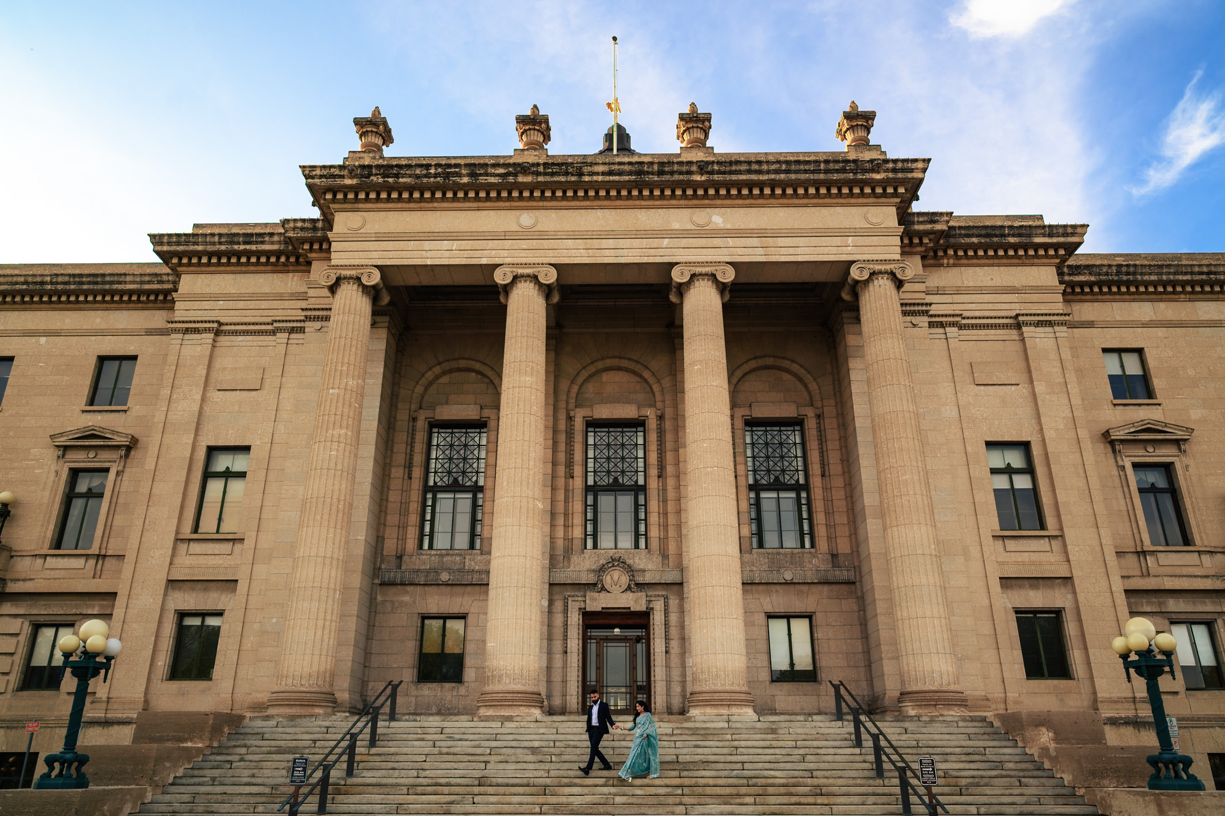 A wide-angle view of a grand neoclassical building with large columns and intricate details sets the stage for an Indian engagement shoot. A couple is visible on the steps, the man wearing a suit and the woman in a formal dress. The sky is partly cloudy, adding contrast to the scene.