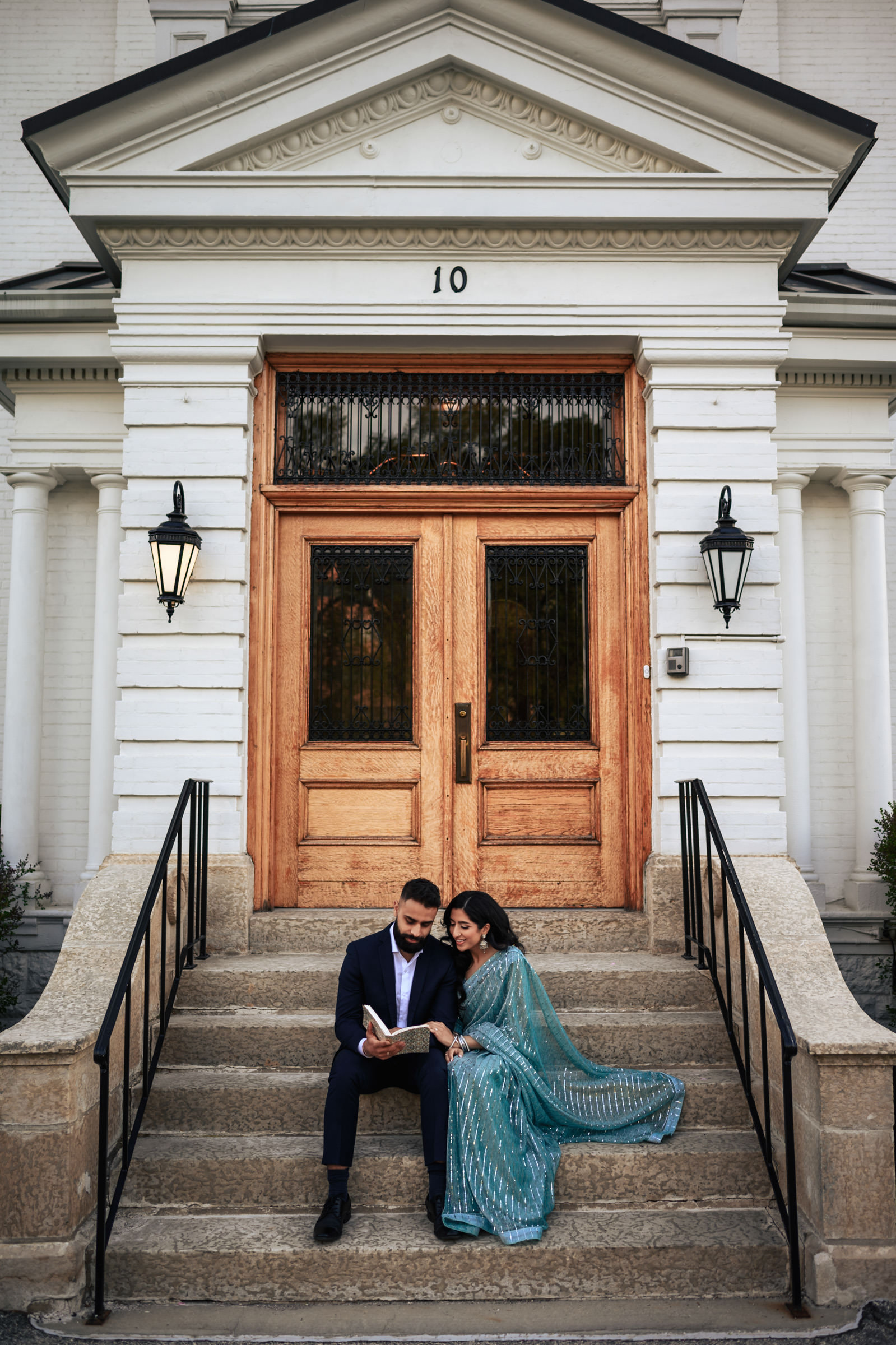 A man in a dark suit and a woman in a teal dress sit together on the steps of a building with large wooden doors and the number 10 above them. They appear to be looking at a book, with the woman resting her head on the man's shoulder, capturing their candid joy in an Indian engagement shoot.