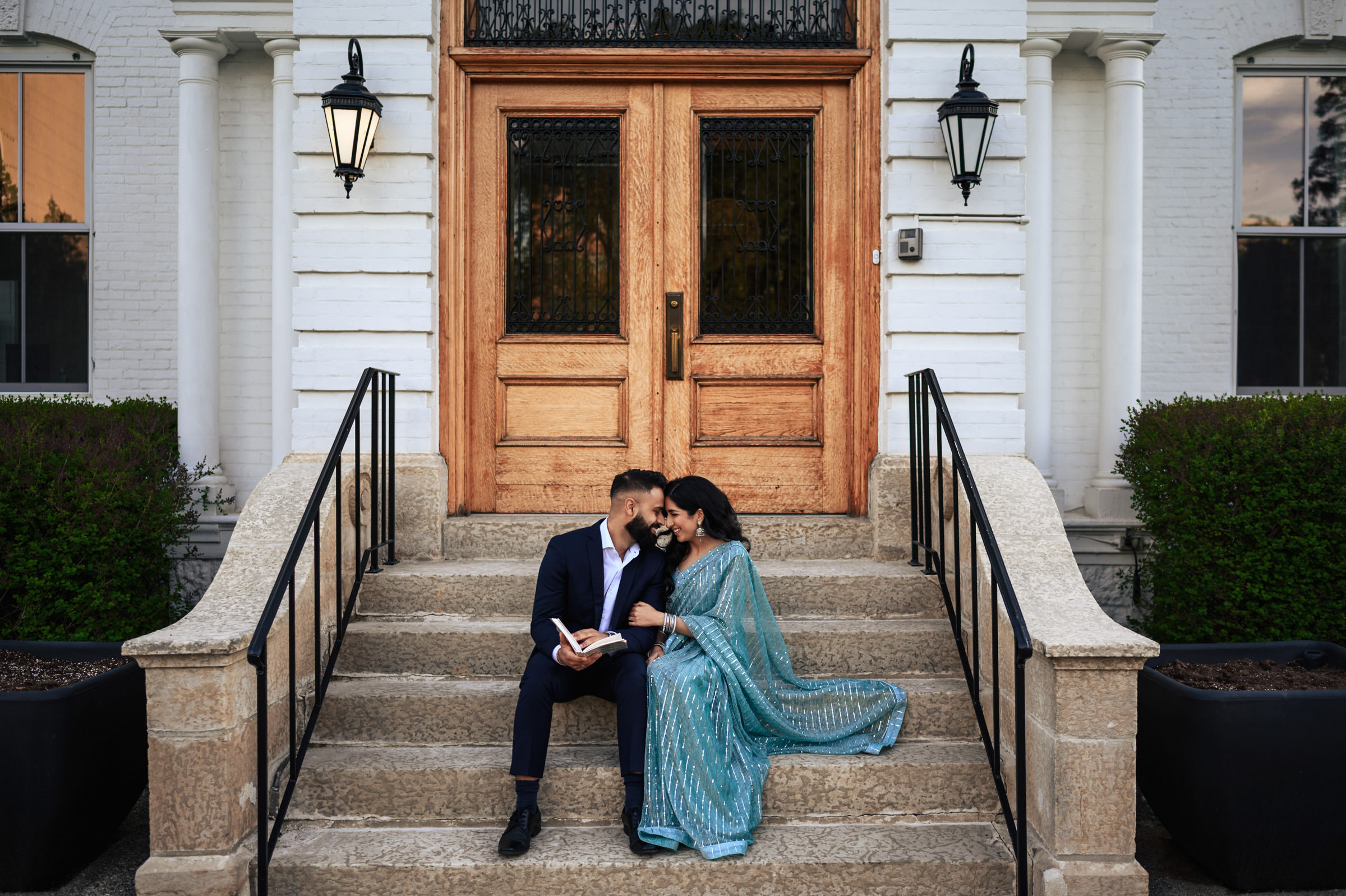A couple sits on the steps of a grand building with wooden doors and black railings. The man, in a dark suit, is reading a book while the woman, in a blue sari, leans in close to him, smiling. Bushes flank the steps, and ornate lamps adorn the entrance—a perfect Indian engagement shoot setting.