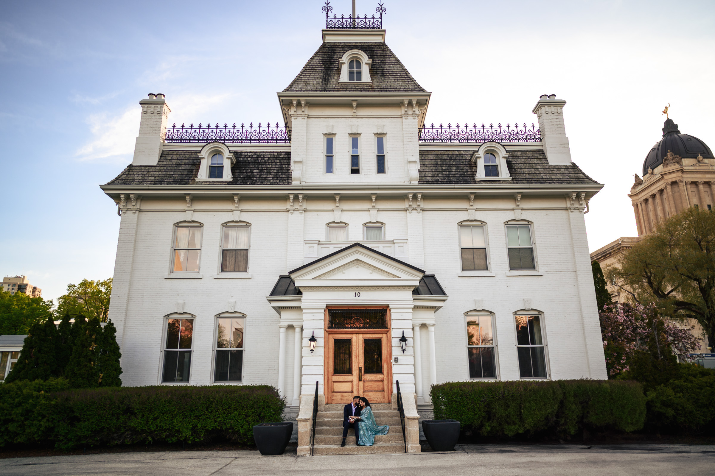 A couple sits on the steps of a large, ornate, white mansion with a mansard roof and intricate ironwork, surrounded by well-kept bushes. The sky is clear with a soft, warm glow—a perfect setting for their Indian engagement shoot. A domed building is visible in the background to the right.