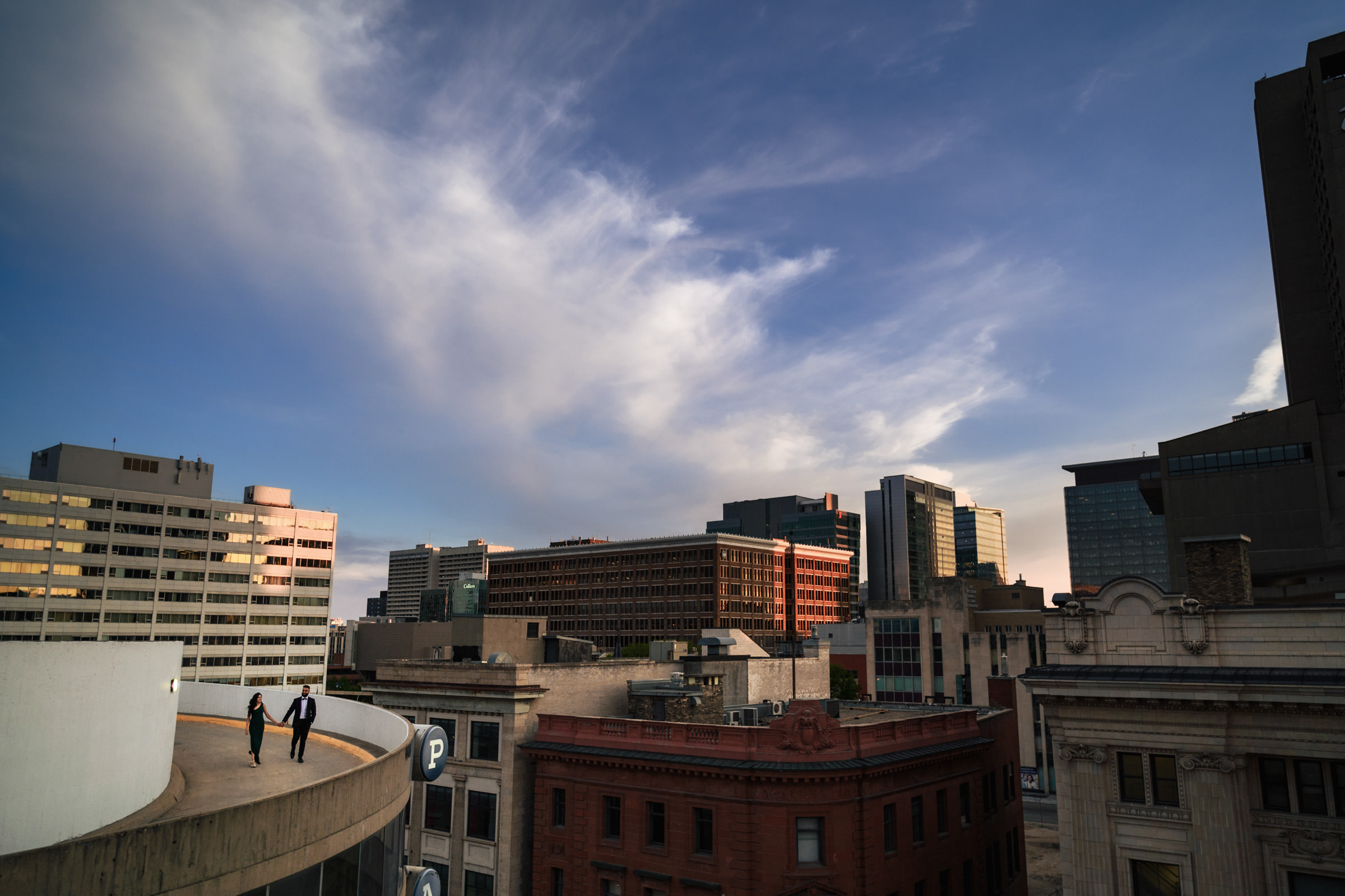 Two people stand on the curved edge of a rooftop parking structure, overlooking a cityscape with modern and older buildings. The sky is clear with a few scattered clouds, and the sun casts a warm light on the scene, highlighting the architectural details—perfect for an Indian engagement shoot.