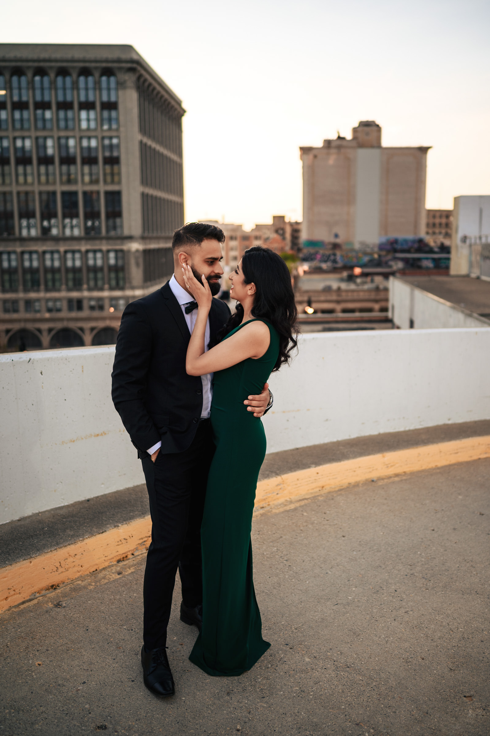A couple stands on an urban rooftop during their Indian engagement shoot. The man, in a dark suit, and the woman, in a green dress, share an intimate moment. The woman gently touches the man's face as both smile warmly. Tall buildings and a clear sky form the beautiful backdrop.
