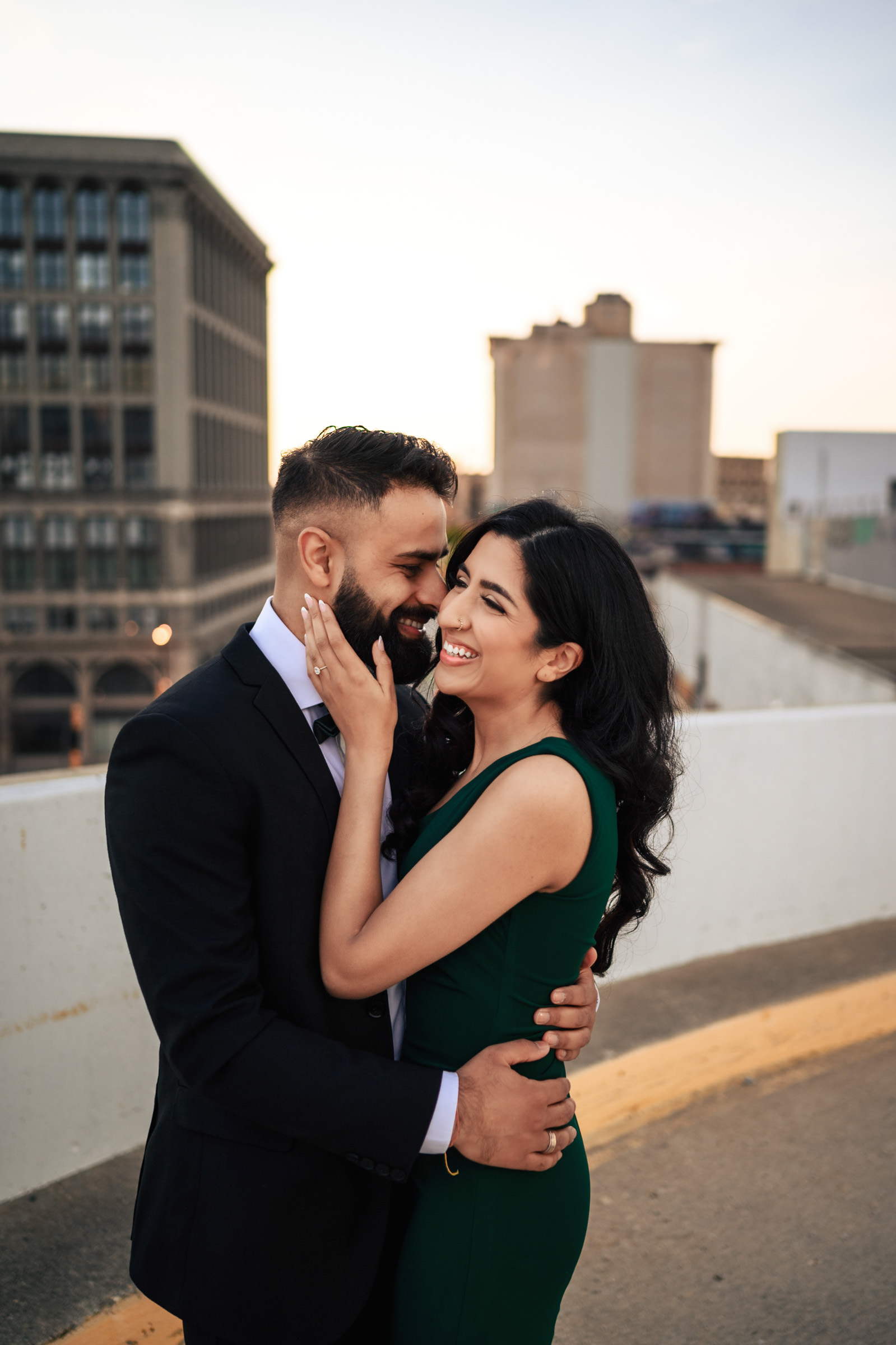 A couple stands on a rooftop parking lot at sunset during their Indian engagement shoot. The man, dressed in a dark suit, clasps the waist of the woman, who is wearing a dark green dress. They smile warmly as she gently touches his face, with urban buildings visible in the background.