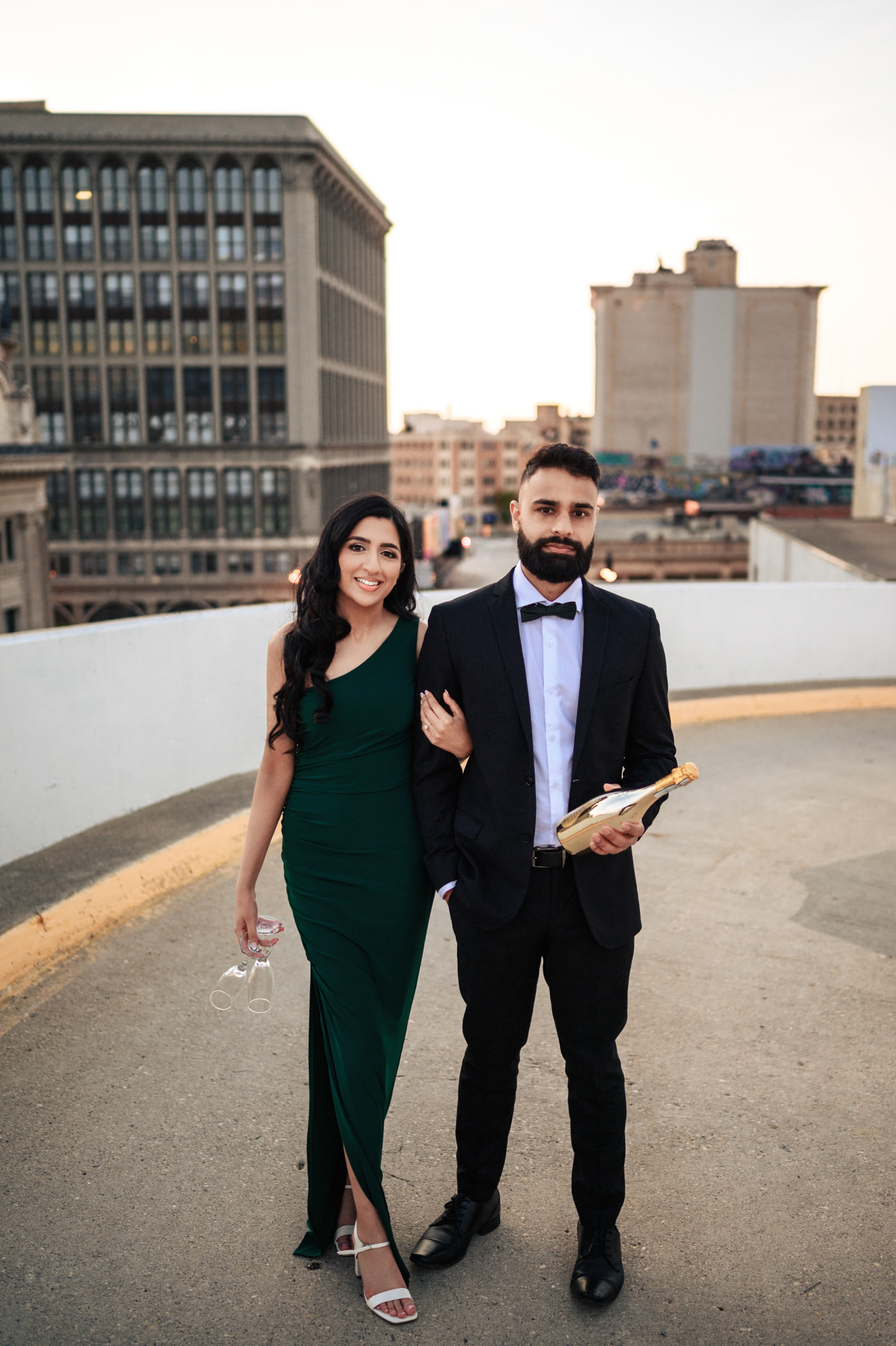 A couple stands on a rooftop at sunset. The woman, in a one-shoulder dark green dress and holding high heels, and the man, dressed in a black suit with a bowtie, hold a bottle of sparkling wine. They smile and pose in front of an urban cityscape, capturing the perfect moment for their Indian engagement shoot.