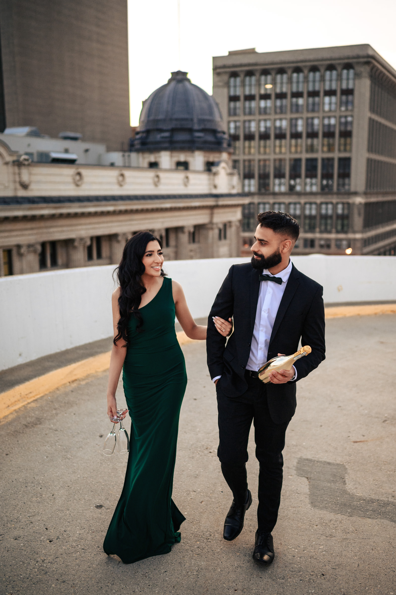 A woman in a green gown and a man in a black suit and bow tie walk arm-in-arm on a rooftop, with city buildings and a domed structure in the background. During their Indian Engagement Shoot, the woman holds a pair of high-heeled shoes, while the man carries a bottle of champagne.