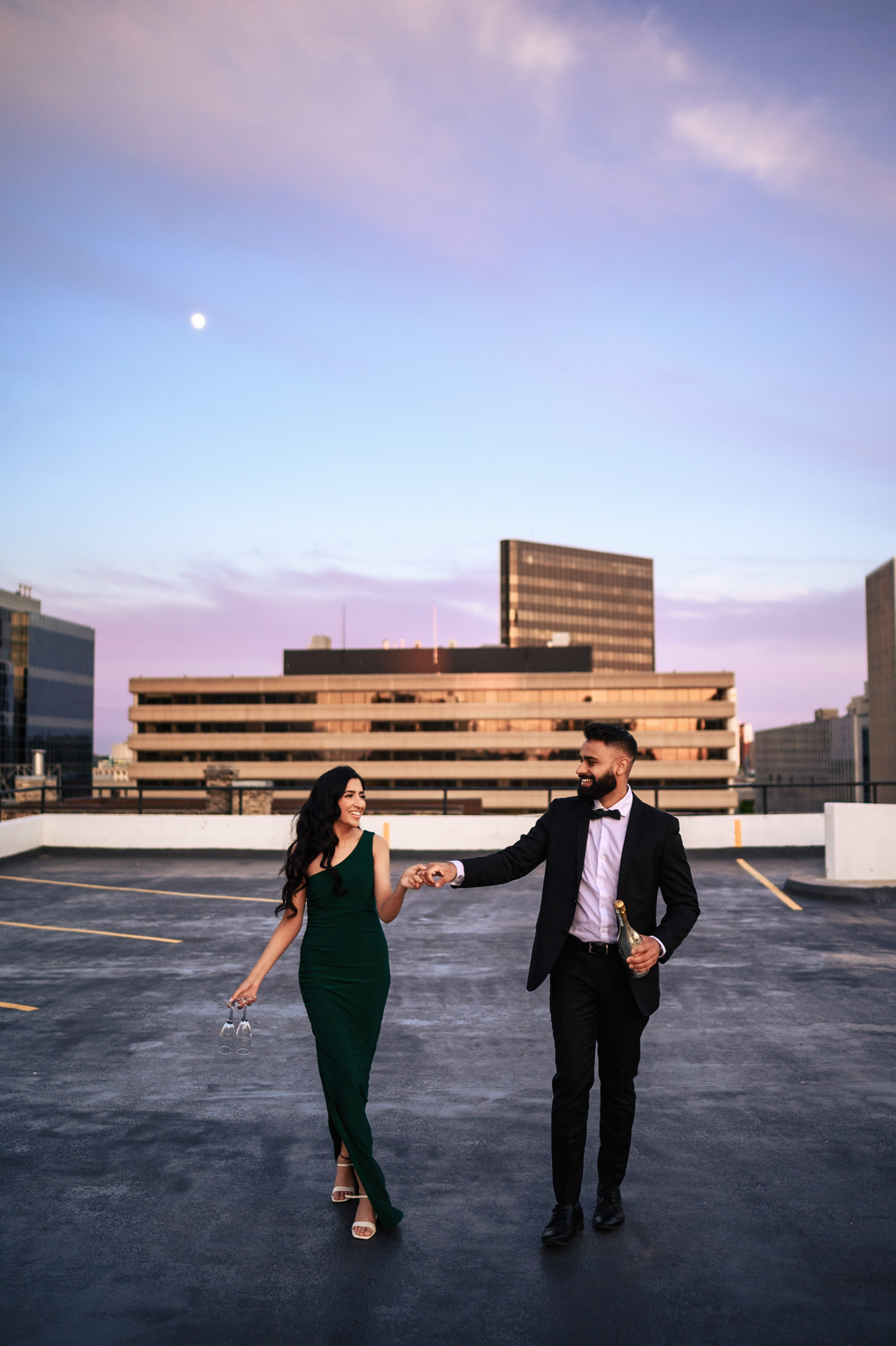 A couple, dressed in formal attire, walks hand in hand on a rooftop parking lot at dusk during their Indian engagement shoot. The woman wears a long green dress and holds high-heeled shoes, while the man in a dark suit holds a drink. They smile at each other with a cityscape in the background.