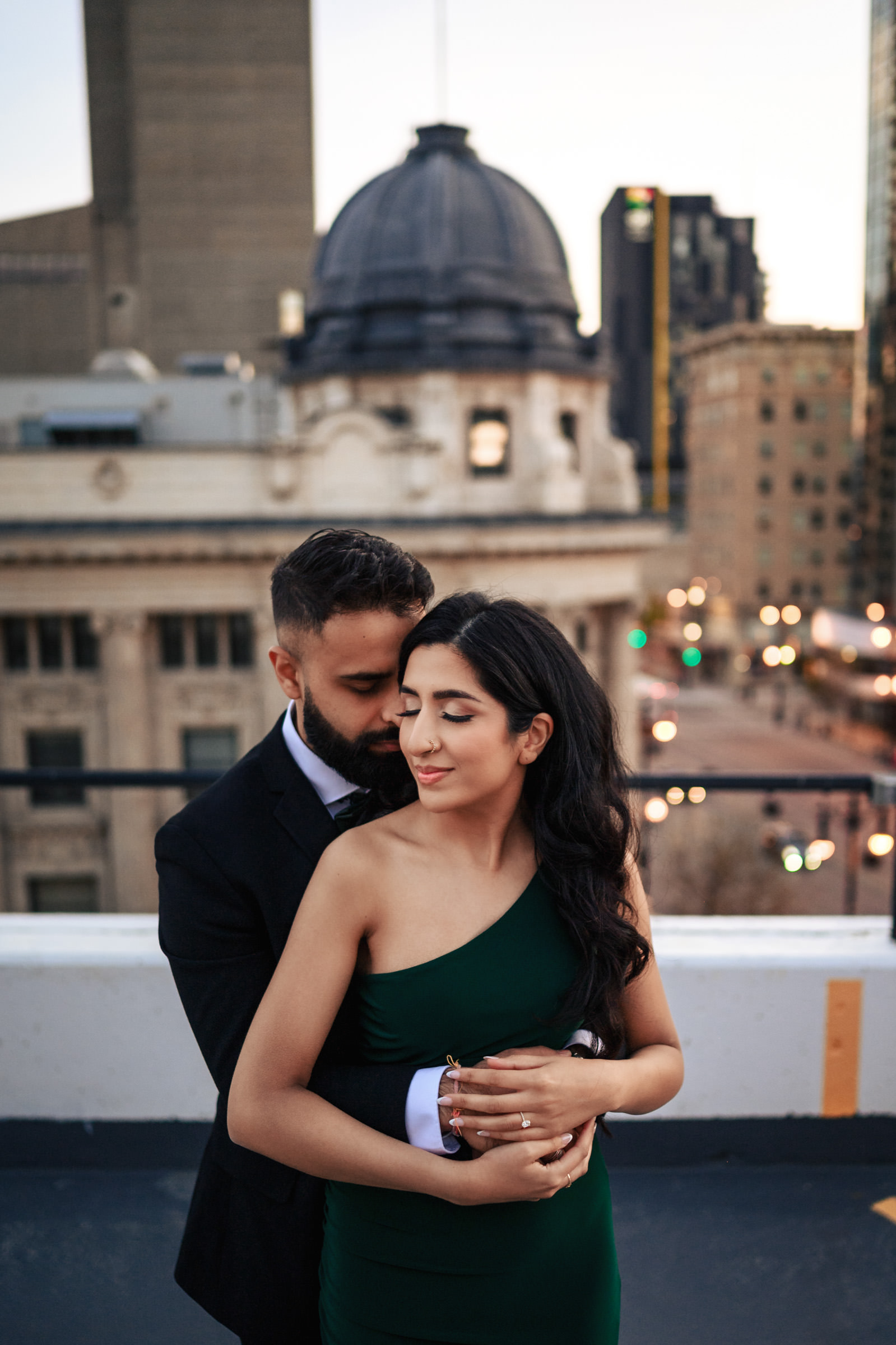 A couple stands on a rooftop with the city's skyline in the background, capturing the essence of an Indian engagement shoot. The man, wearing a dark suit, embraces the woman from behind. She is clad in an elegant, off-the-shoulder green dress. Both have their eyes closed, sharing a tender moment.