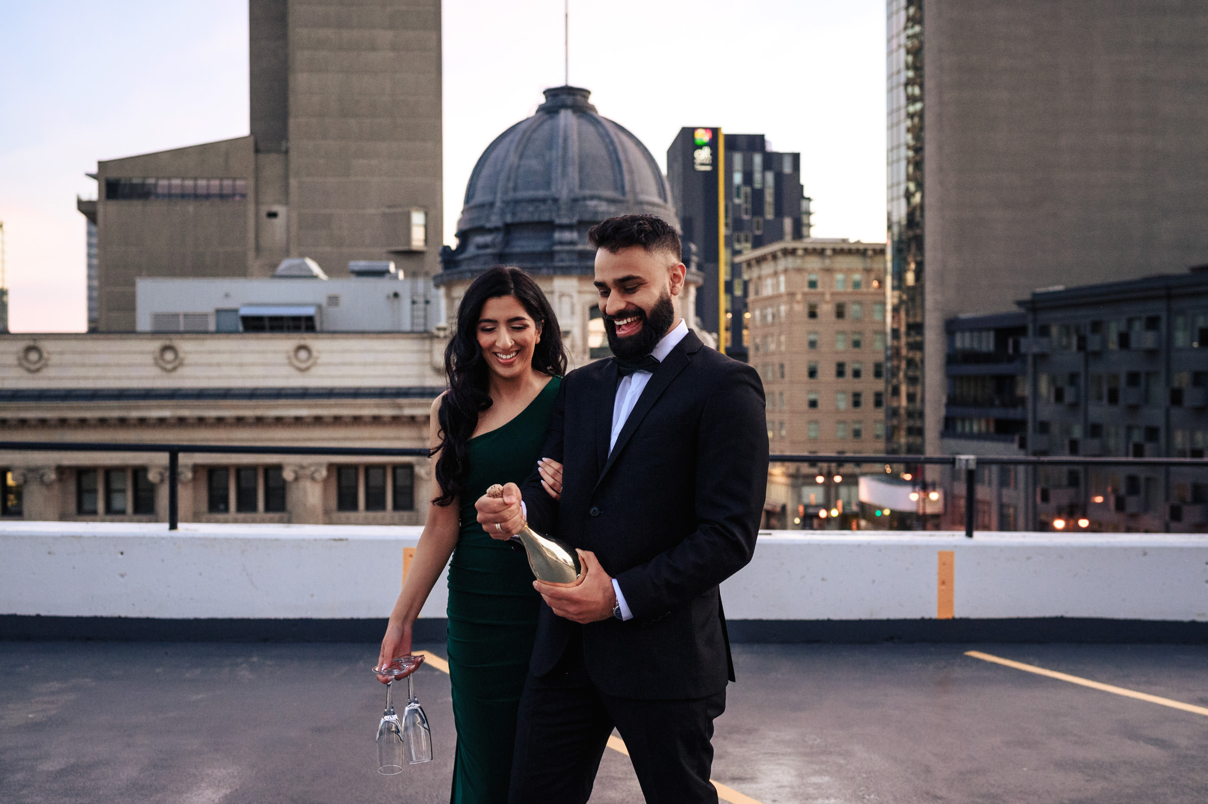A couple stands on a rooftop, with a cityscape in the background. During their Indian engagement shoot, the man, in a suit, opens a bottle of champagne while the woman in a green dress smiles and holds two champagne glasses. They appear to be celebrating with high-rise buildings visible behind them.