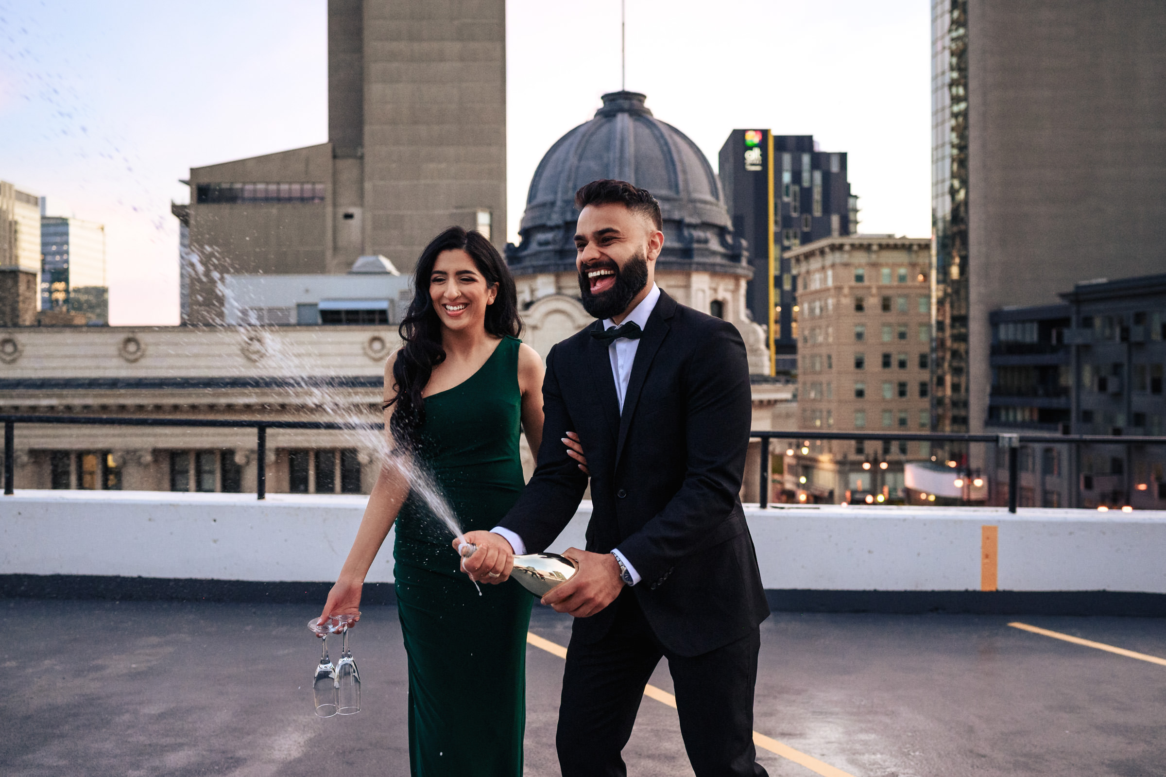 A smiling couple stands on a city rooftop during their Indian engagement shoot, with the man in a black suit and bow tie opening a champagne bottle, spraying its contents. The woman, in a green dress, holds her shoes in one hand and looks on joyfully. Urban buildings and a domed structure are visible in the background.