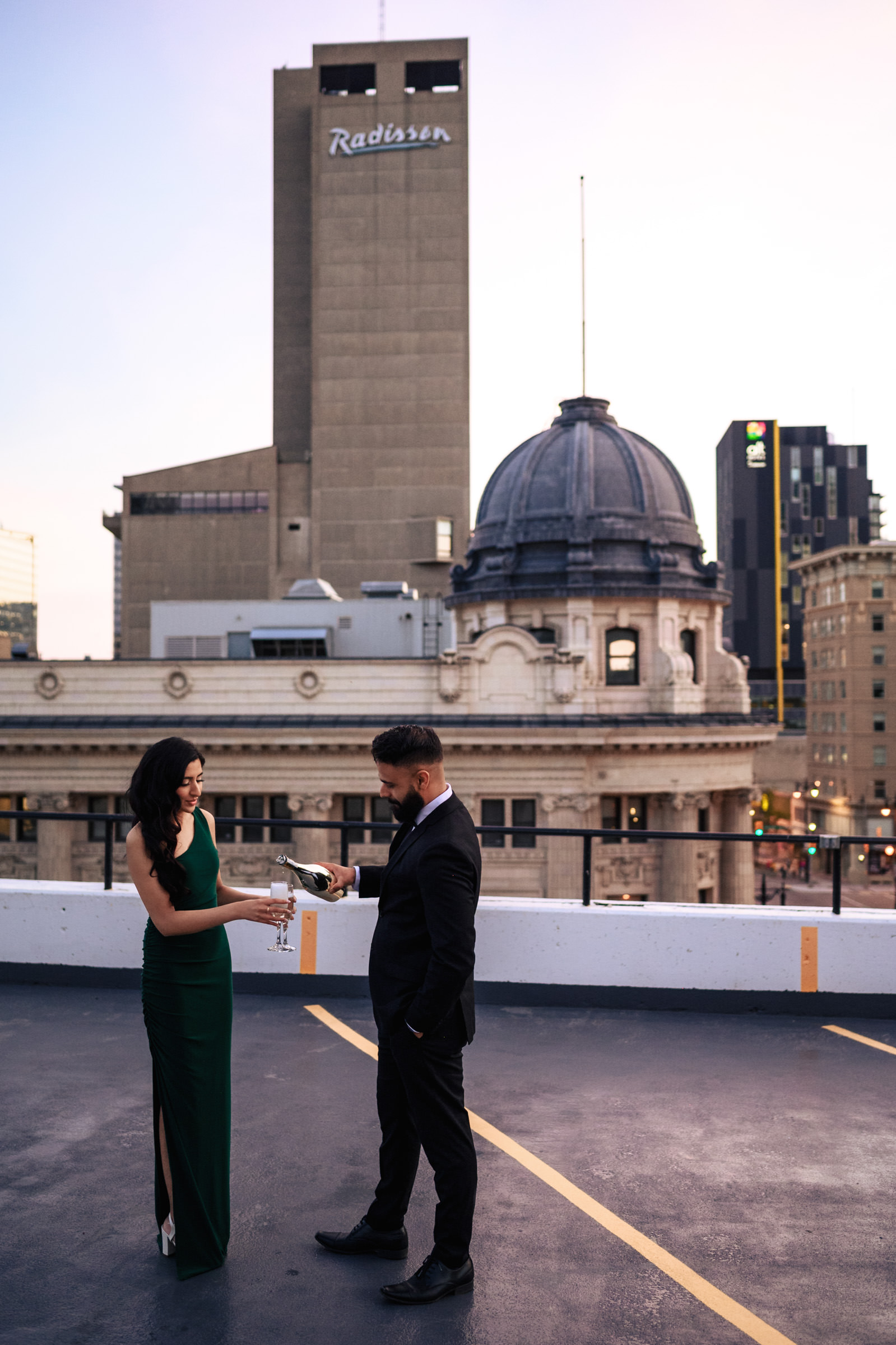 A woman in a green dress and a man in a black suit stand on a rooftop parking lot during their Indian engagement shoot. The woman is pouring champagne into a glass held by the man. Behind them, an old building with a dome and a tall hotel building with the "Radisson" sign are visible.