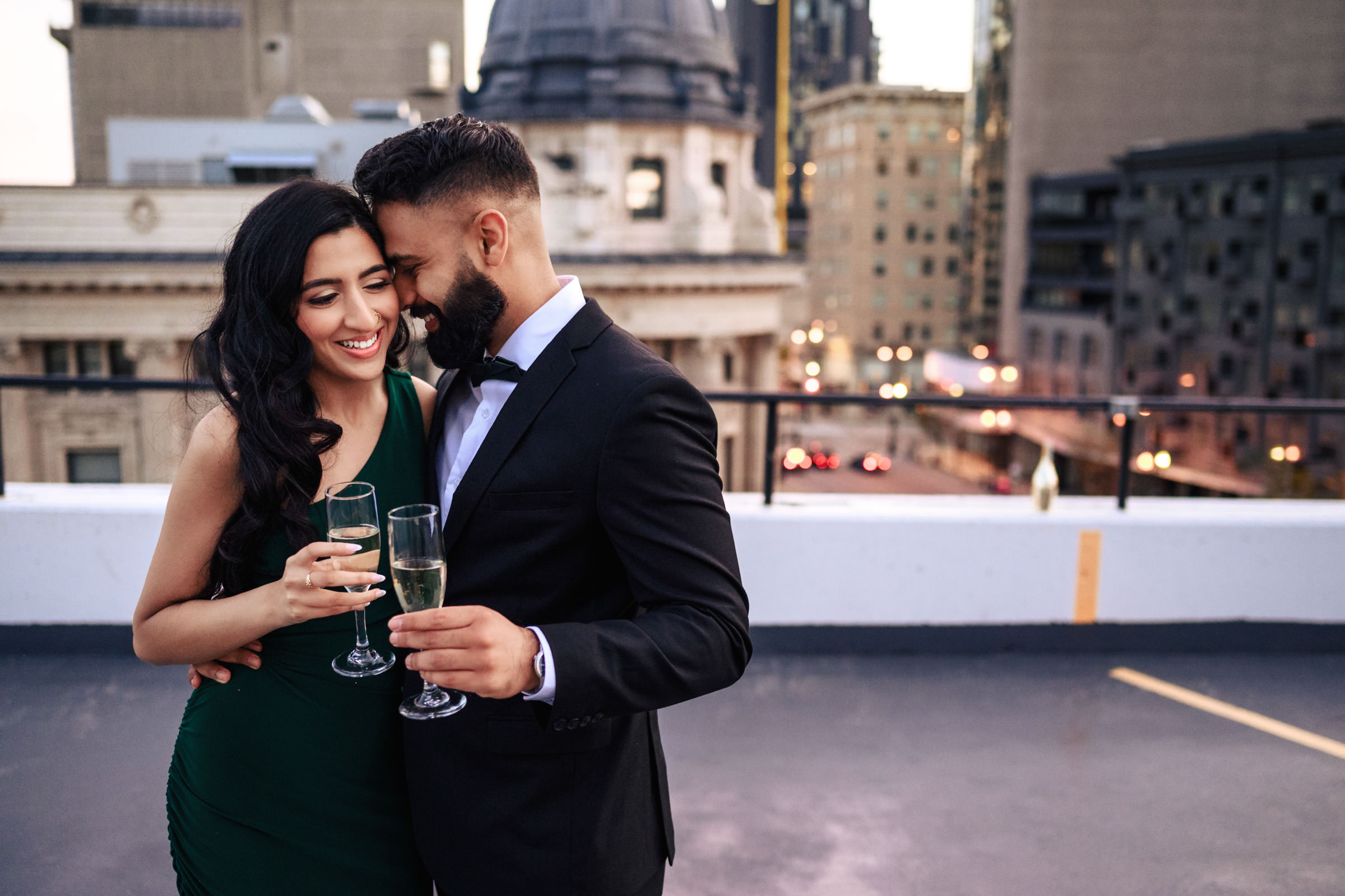 A couple stands close together on a rooftop, smiling and holding glasses of champagne during their Indian engagement shoot. The man is wearing a black suit, while the woman is in a green dress. The cityscape in the background is blurred, showcasing tall buildings and shimmering lights.