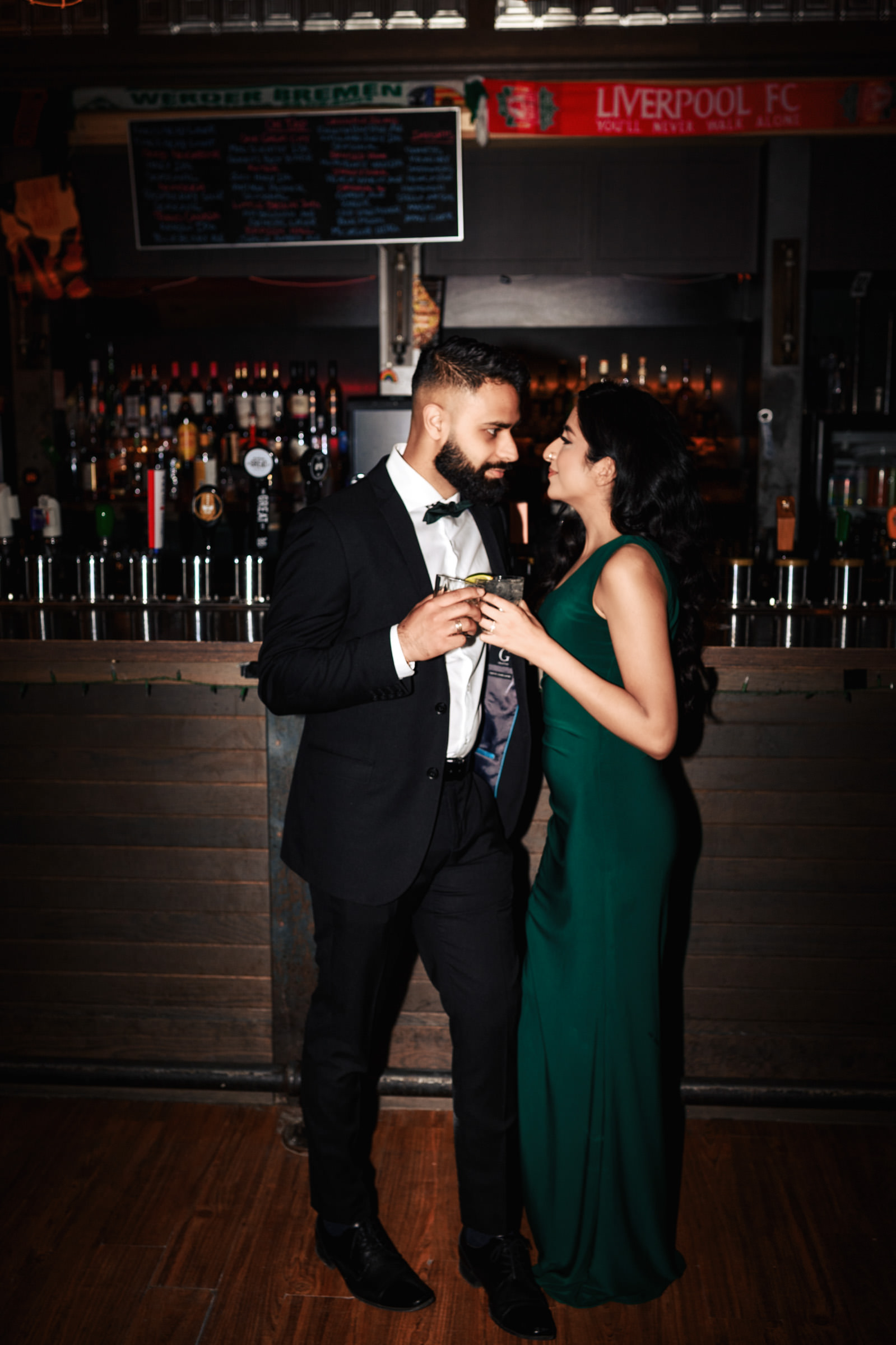 A man in a black tuxedo and a woman in a green dress stand close together, smiling, and holding drinks in a dimly lit bar. The bar counter behind them is lined with various bottles, and a "Liverpool FC" sign is visible above it, capturing the essence of an Indian engagement shoot.