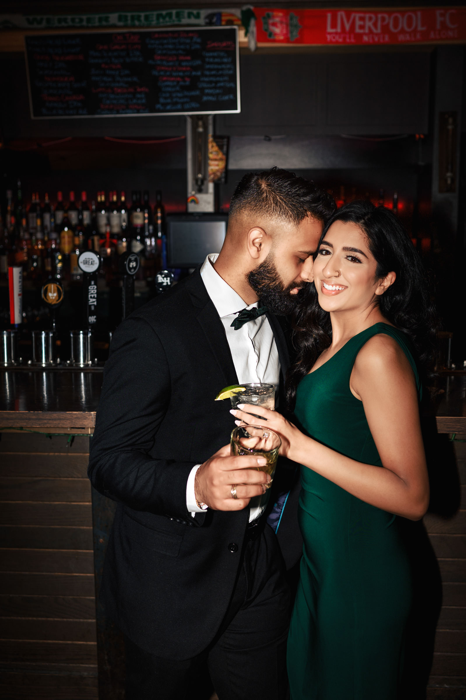 A man in a black suit and bow tie and a woman in a green dress share a happy moment at a bar with drinks in hand. The bar, adorned with a "Liverpool FC" scarf above its well-stocked shelves, sets the perfect backdrop for their Indian engagement shoot as they smile and lean close to each other.